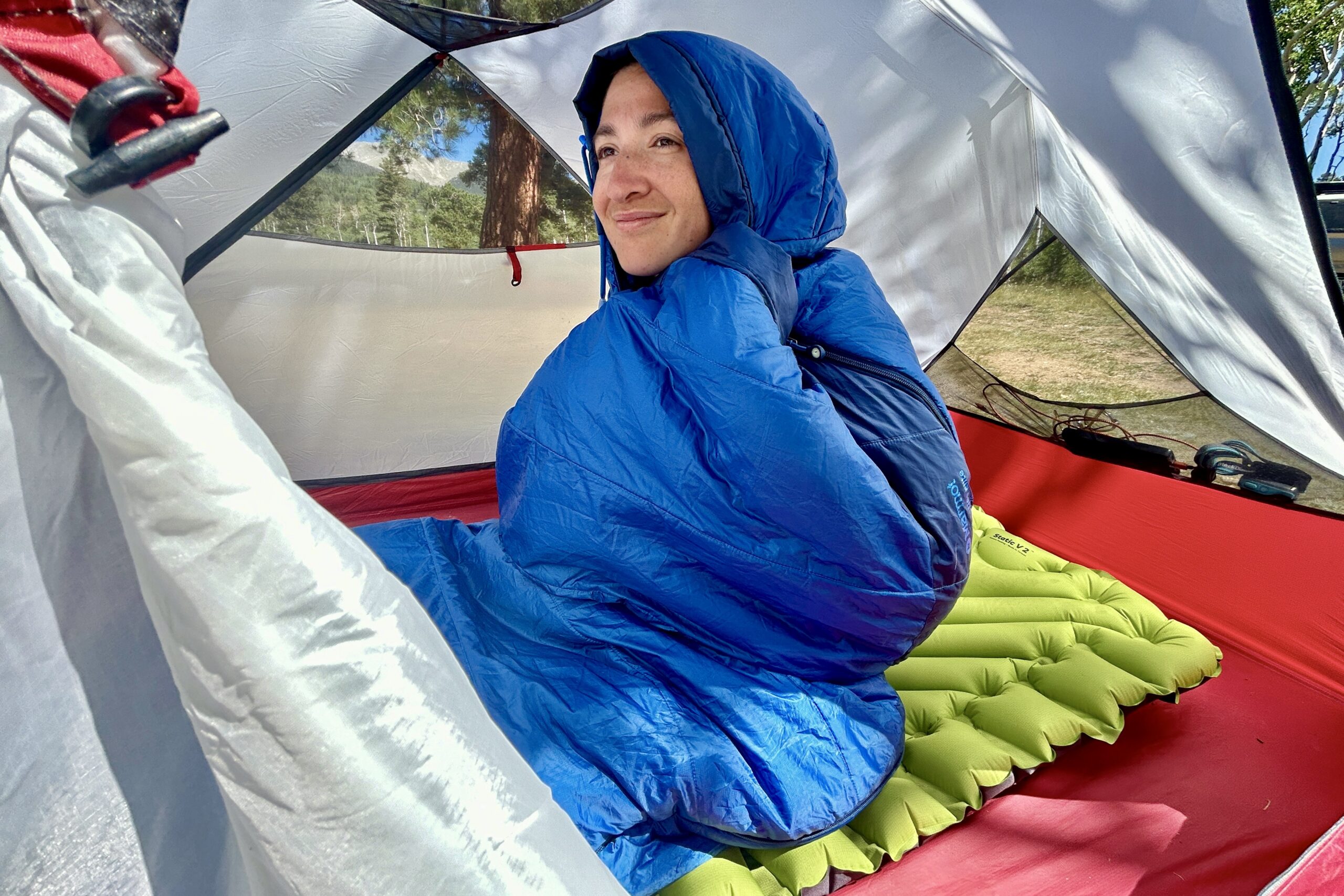 A close-up picture of a woman sitting upright and snuggling into the Marmot Trestles 20 sleeping bag, with the hood up around her face, in a red tent in the sun.