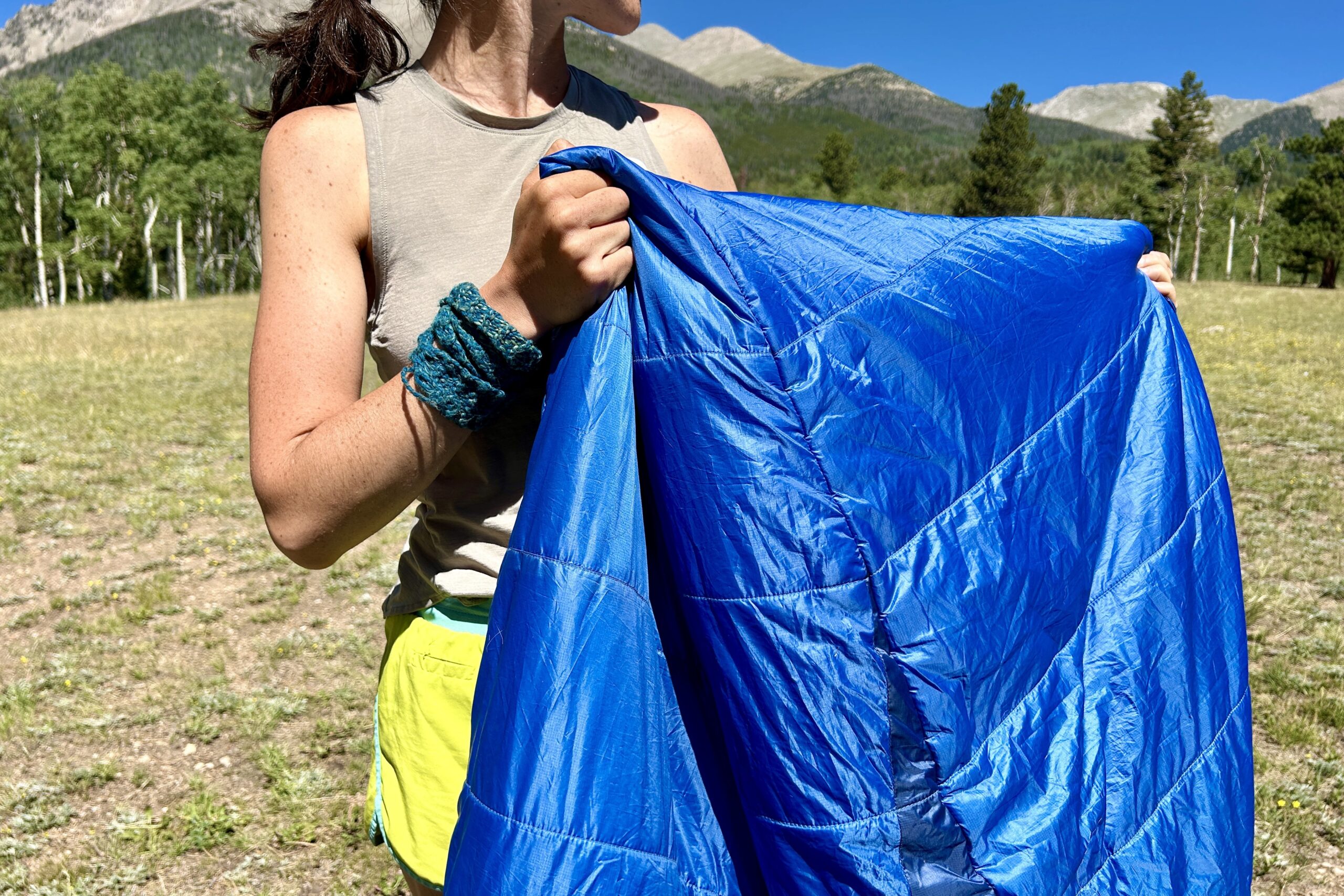 A close-up image of a woman standing outside with mountains in the background, holding up a blue sleeping bag.