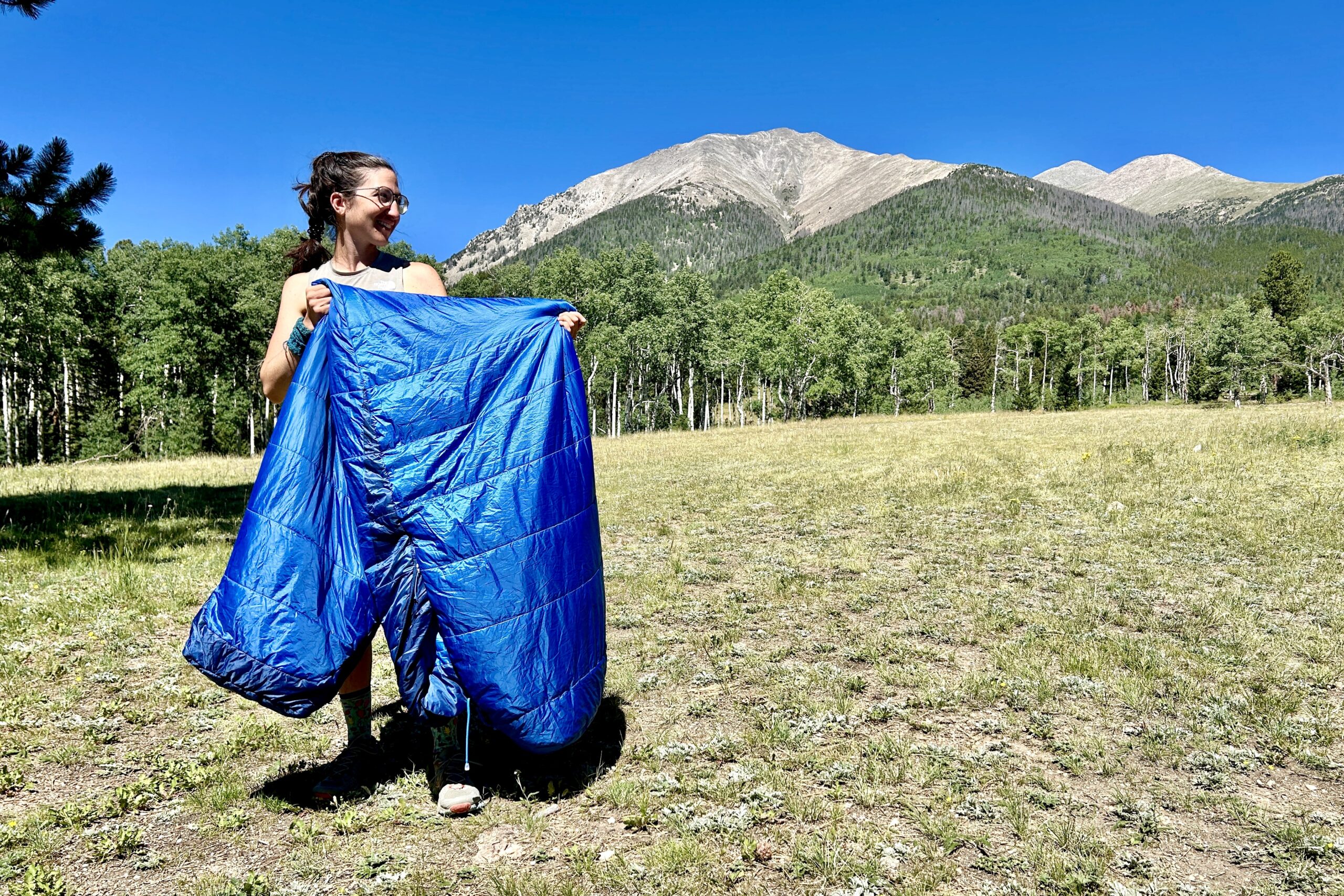 A woman stands outside with mountains in the background, holding up a blue sleeping bag, while she looks off int he distance.