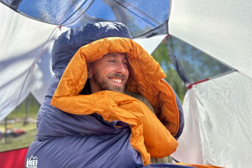 A man sits up in a sleeping bag with the hood over his head. He is smiling and looking off camera.