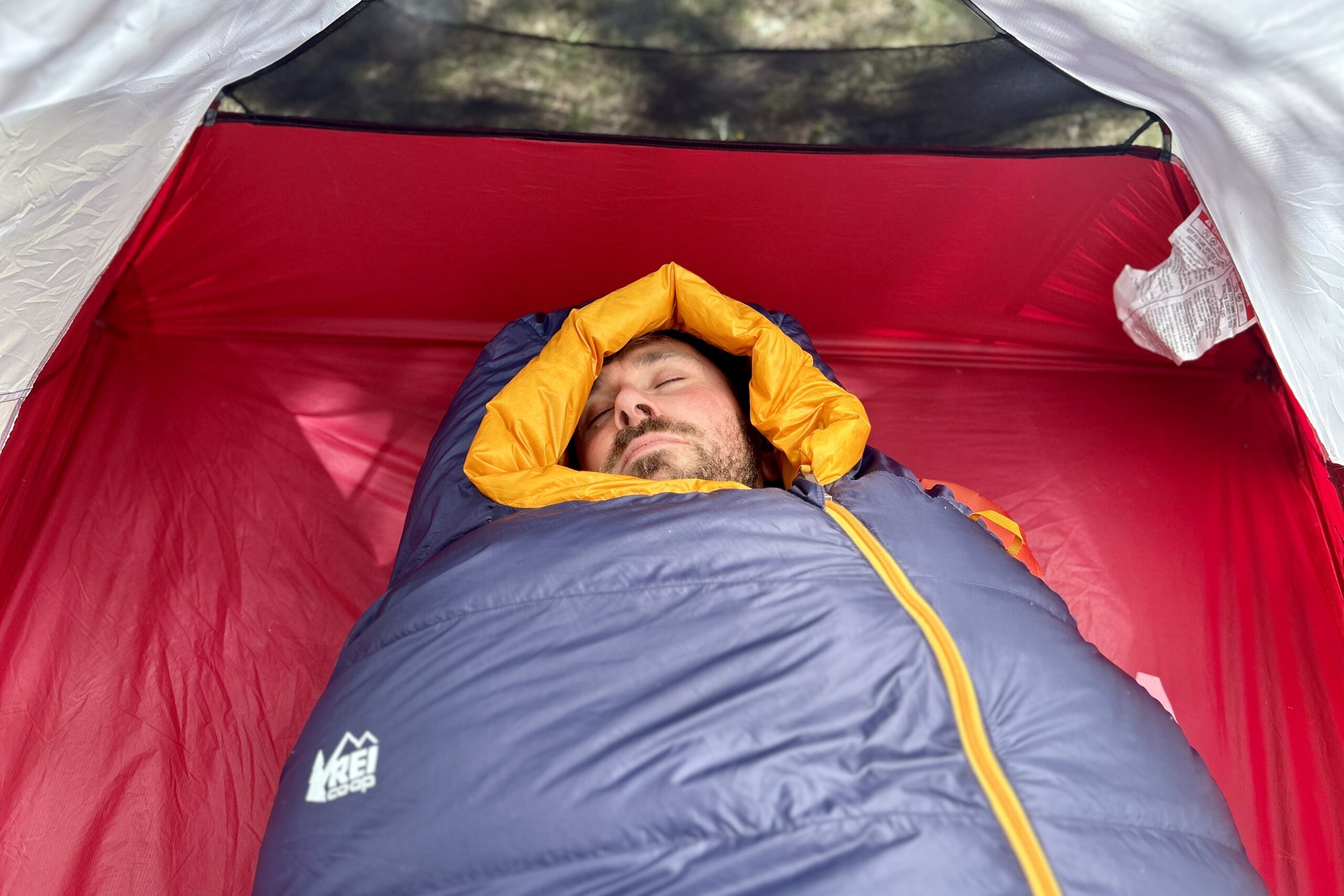 A man in a sleeping bag rests with the mummy hood cinched around his face in a tent.