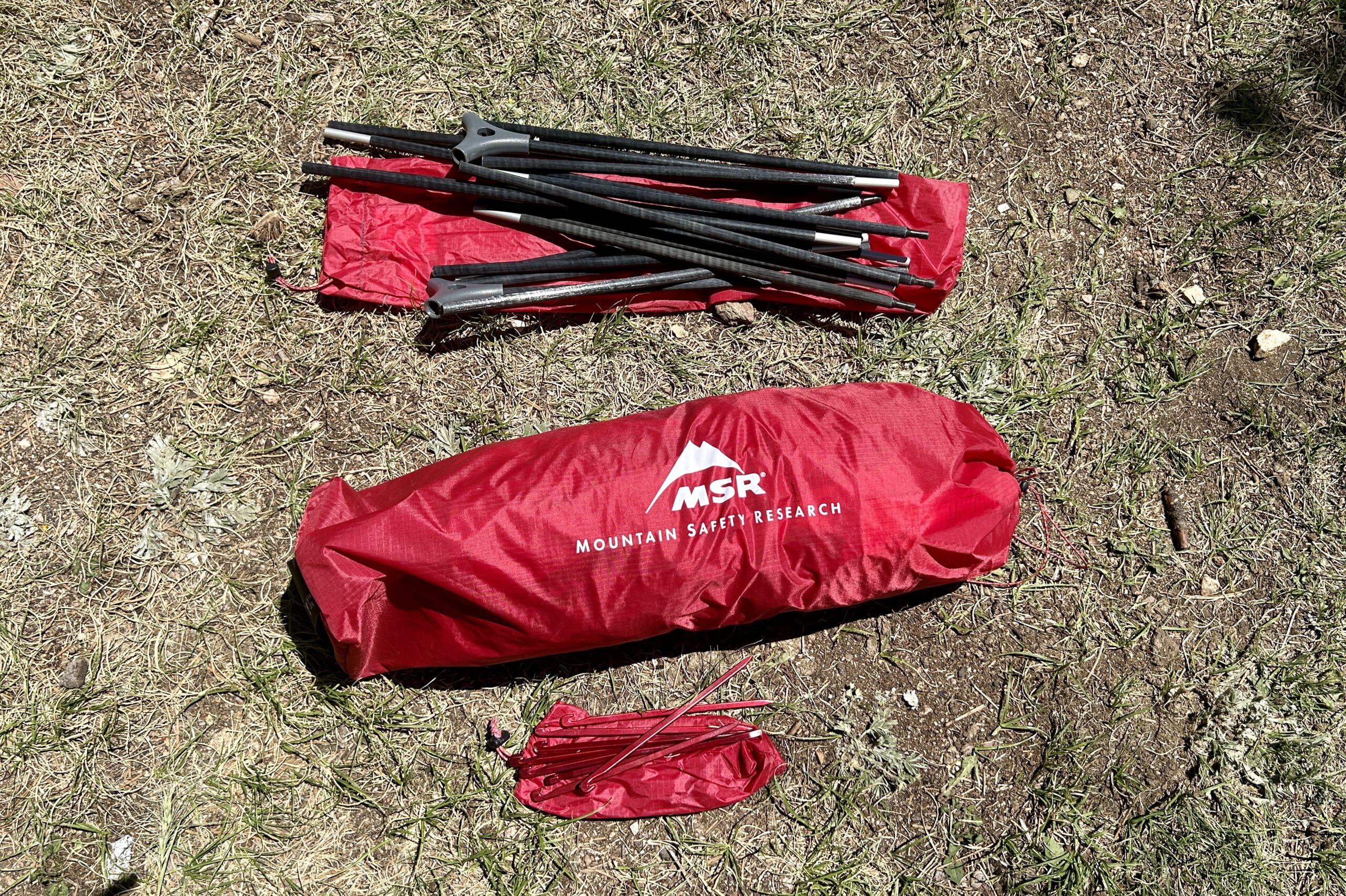 A tent stuff sack, poles, and stakes rest on the ground. next to each other.