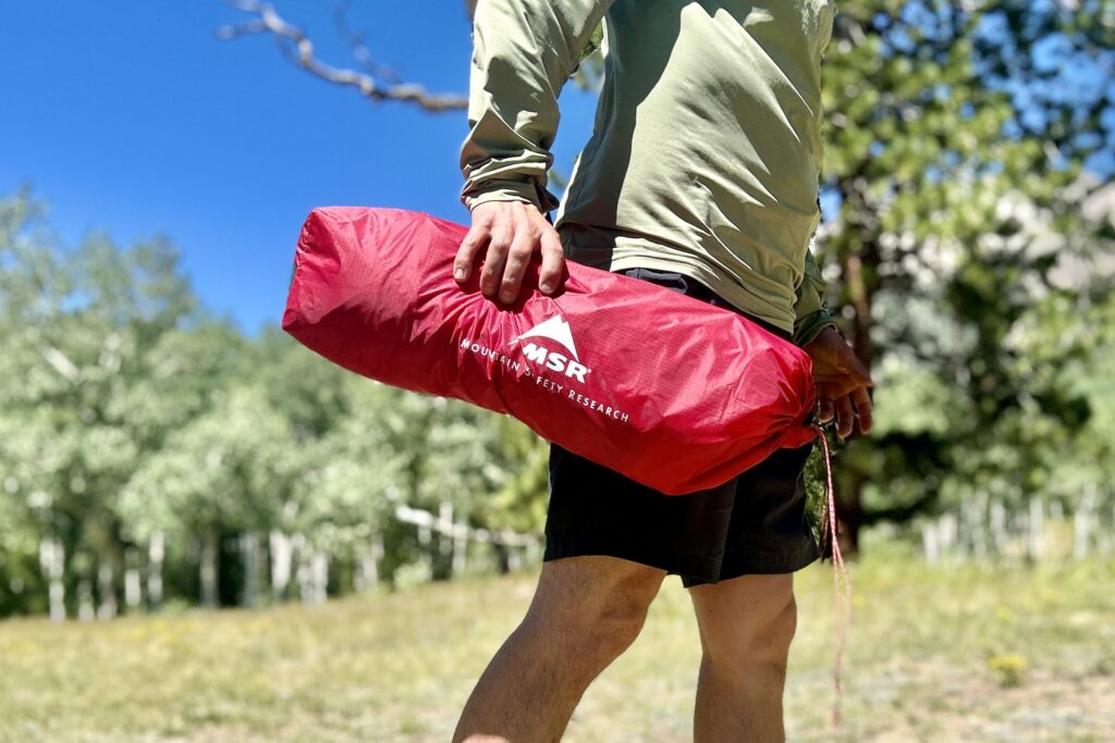 A man walks through the woods carrying a bright red tent stuff bag