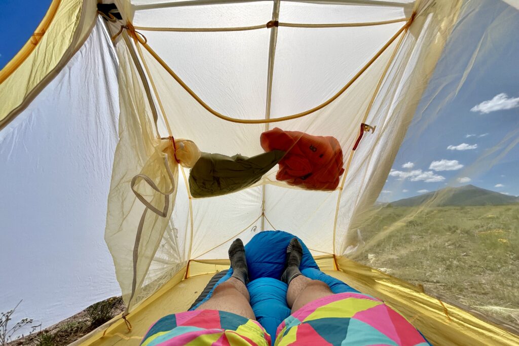 A person lays in a tent on a sunny day with some objects in the overhead pocket.