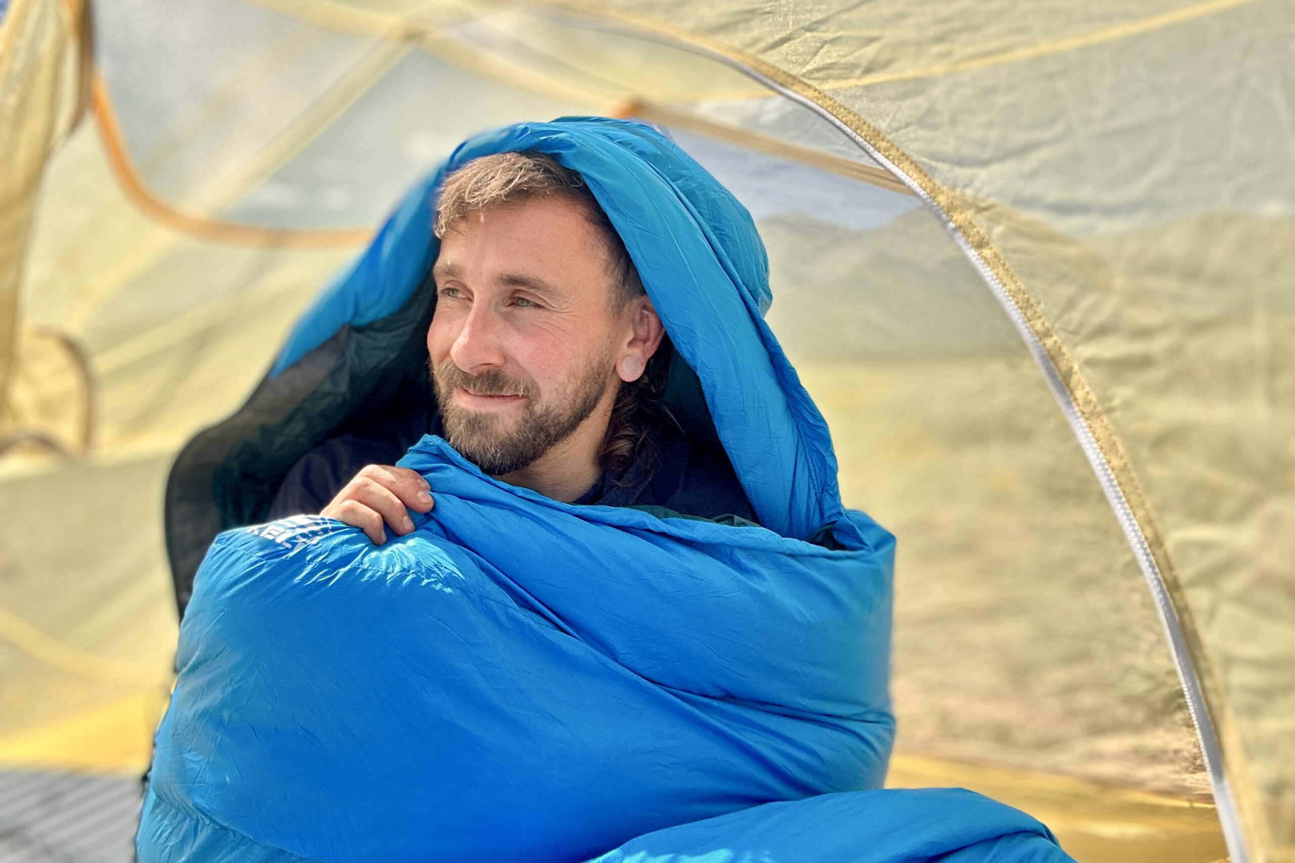 A close up image of a man smiling and looking off in the distance with a sleeping bag around his shoulders, sitting up in his tent, as the morning sun streams through.