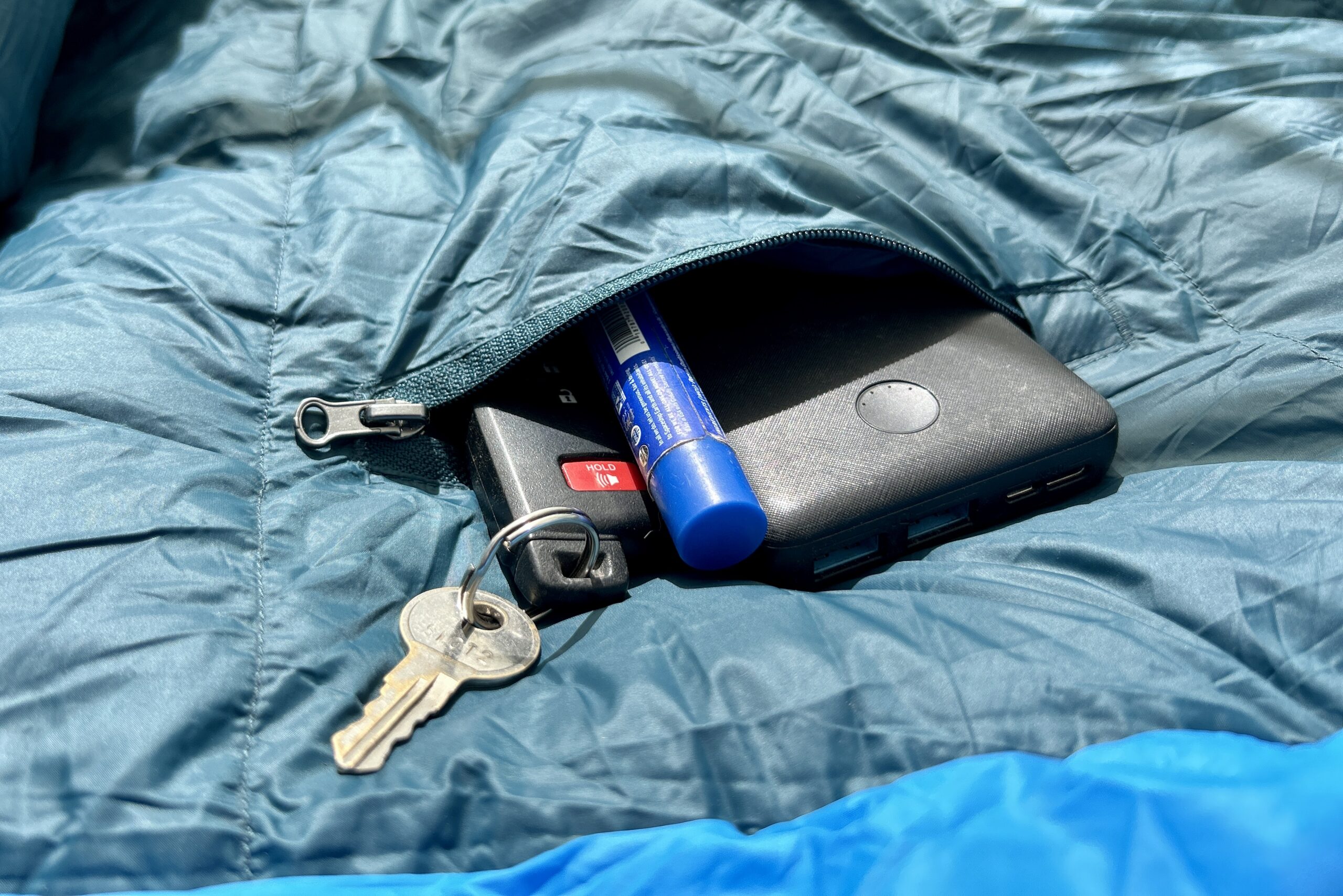 A close up image of keys, a chapstick, and a battery pack sticking out of a zippered internal sleeping bag pocket.