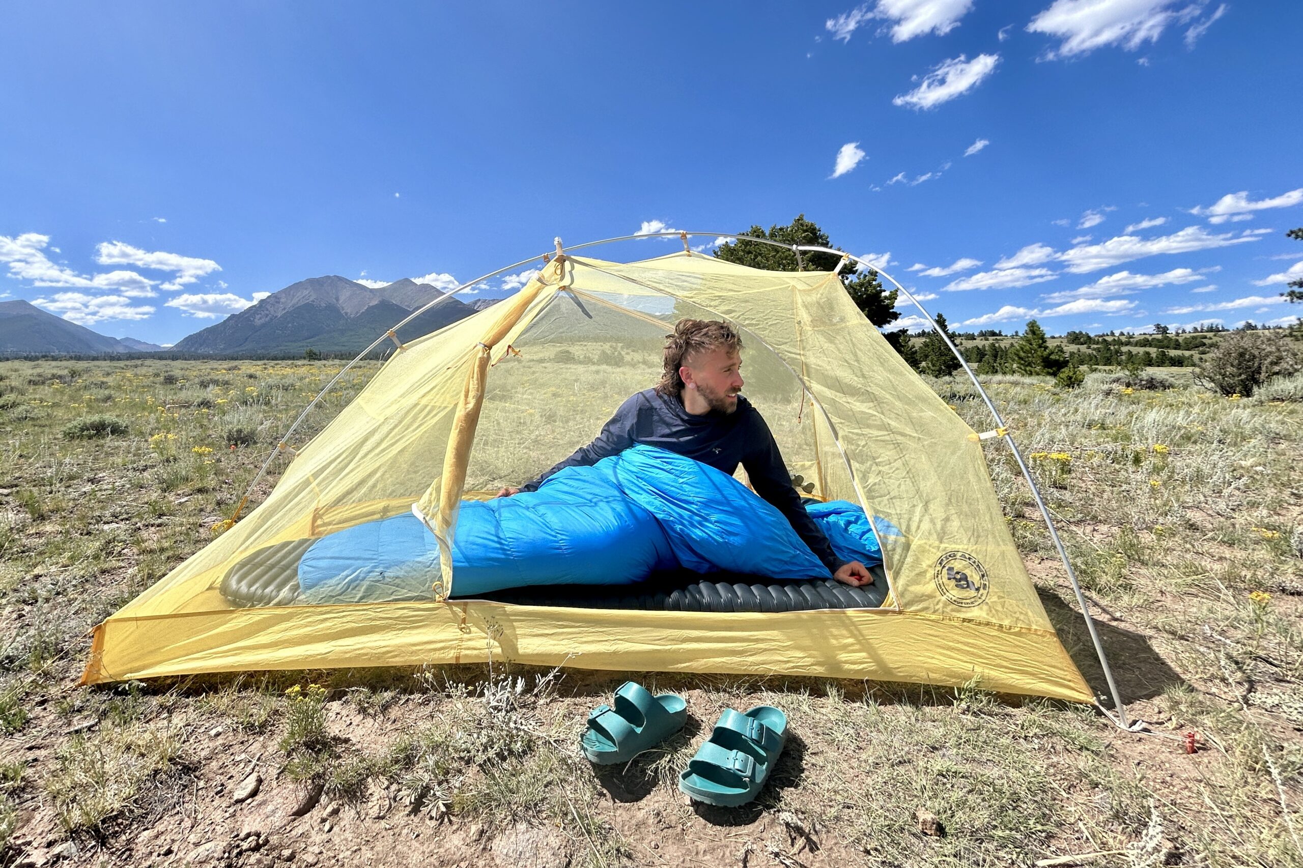 A man sits in a sleeping bag, on a pad, in a tent, looking off in the background. He is camped in a wide field with large mountains in the backgorund.