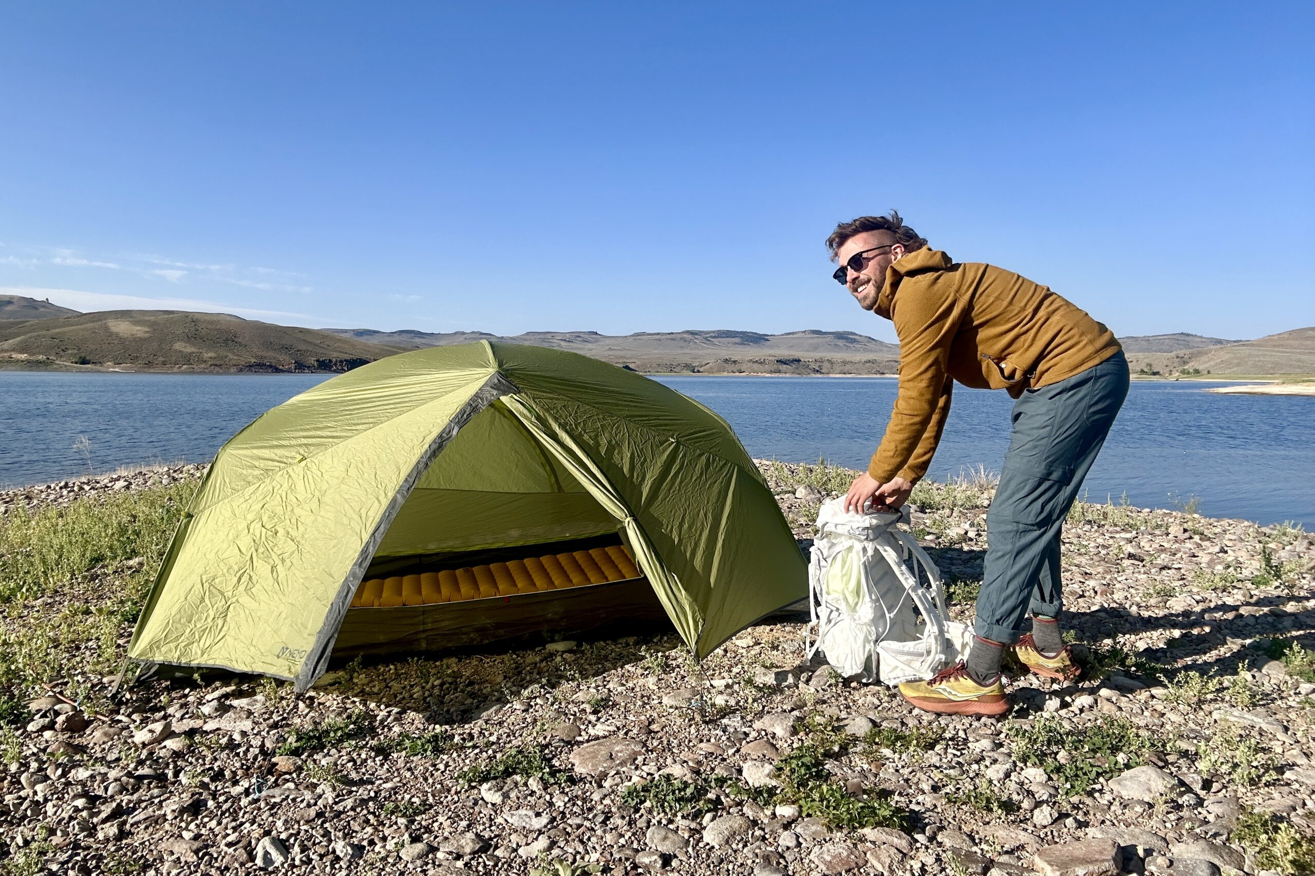 A man unpacks a backpack in front of a tent and there is a lake in the background.