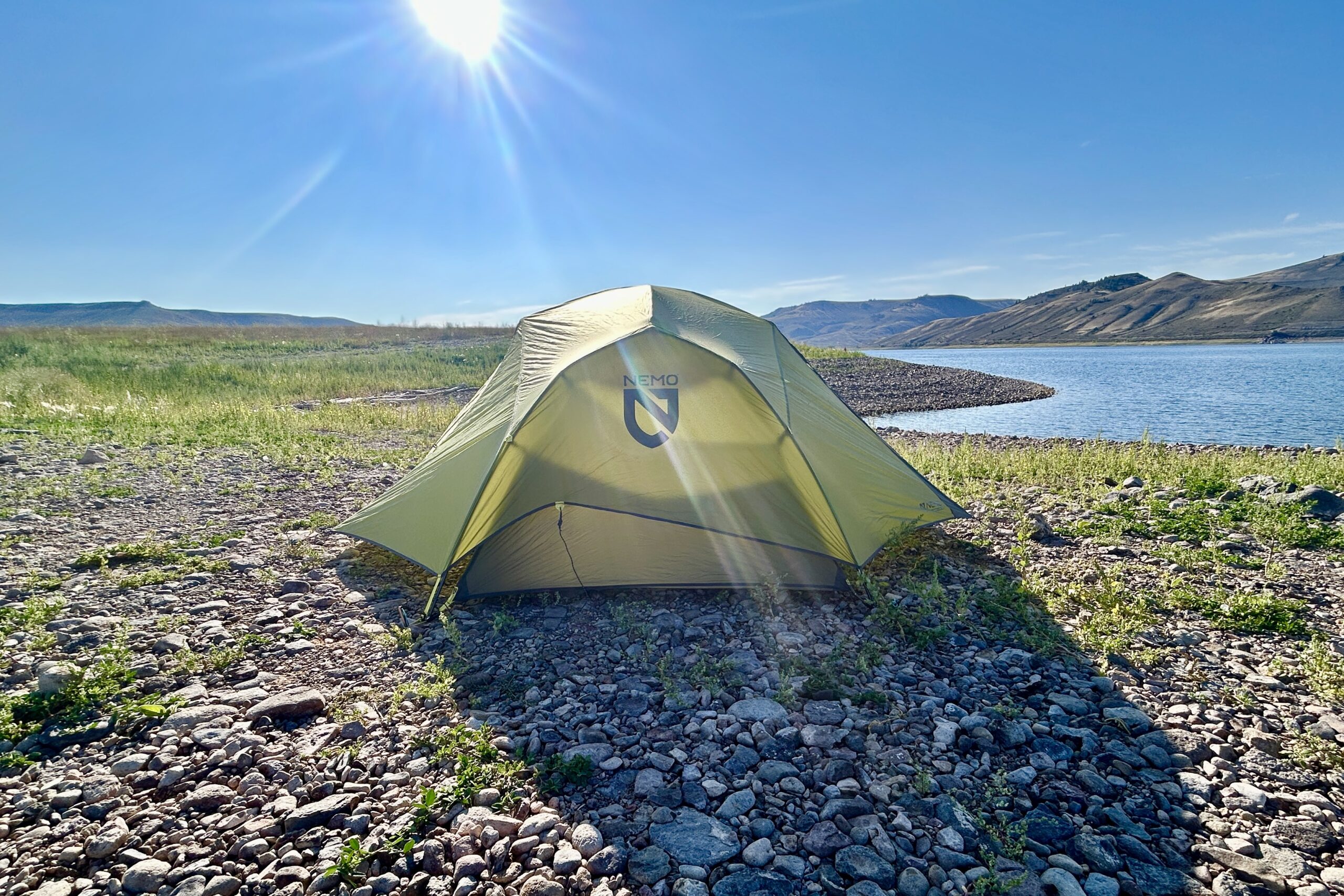 A tent stands alone in the center of a rocky beach with a lake in the background.