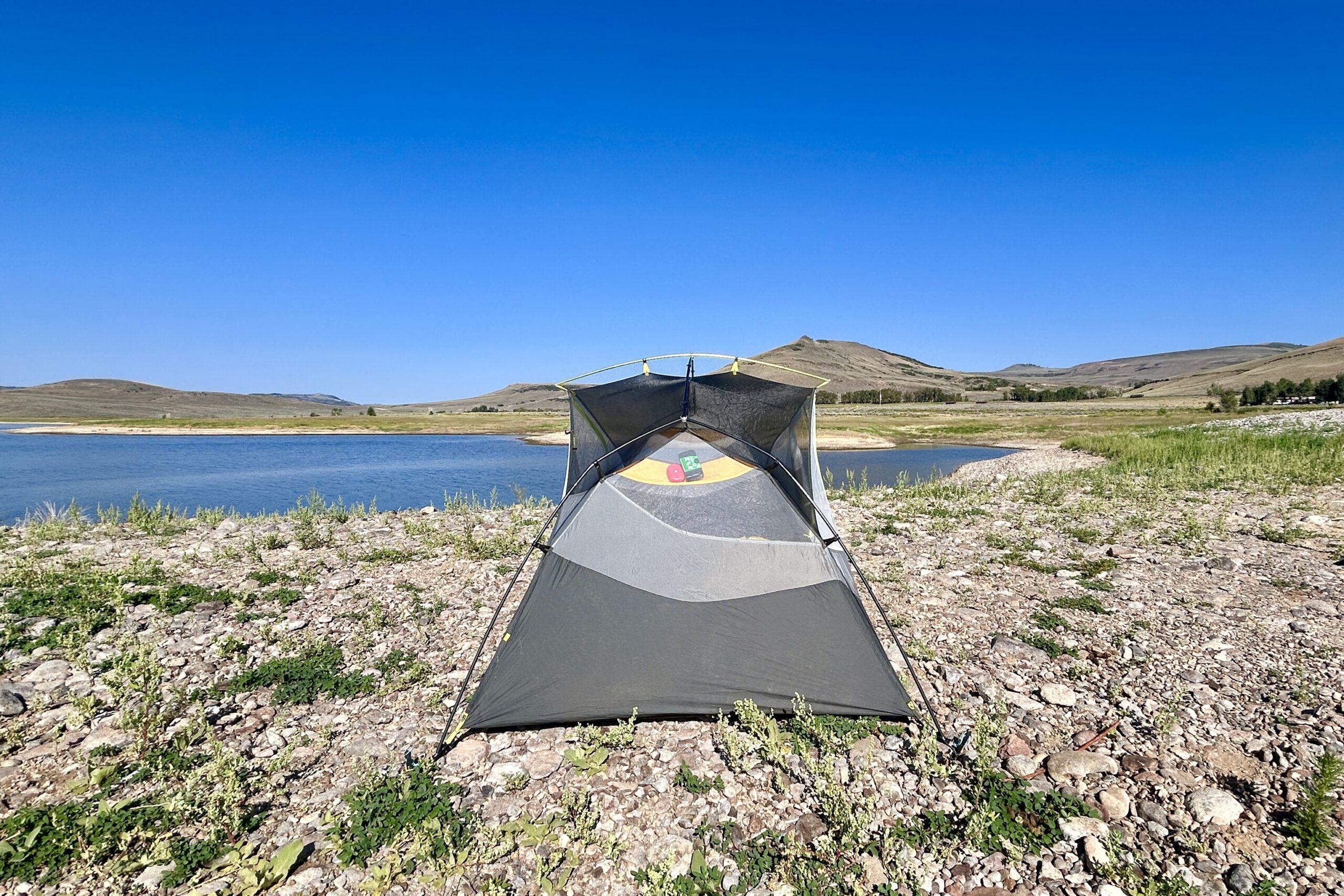 A tent stands alone in the center of a rocky beach with a lake in the background.