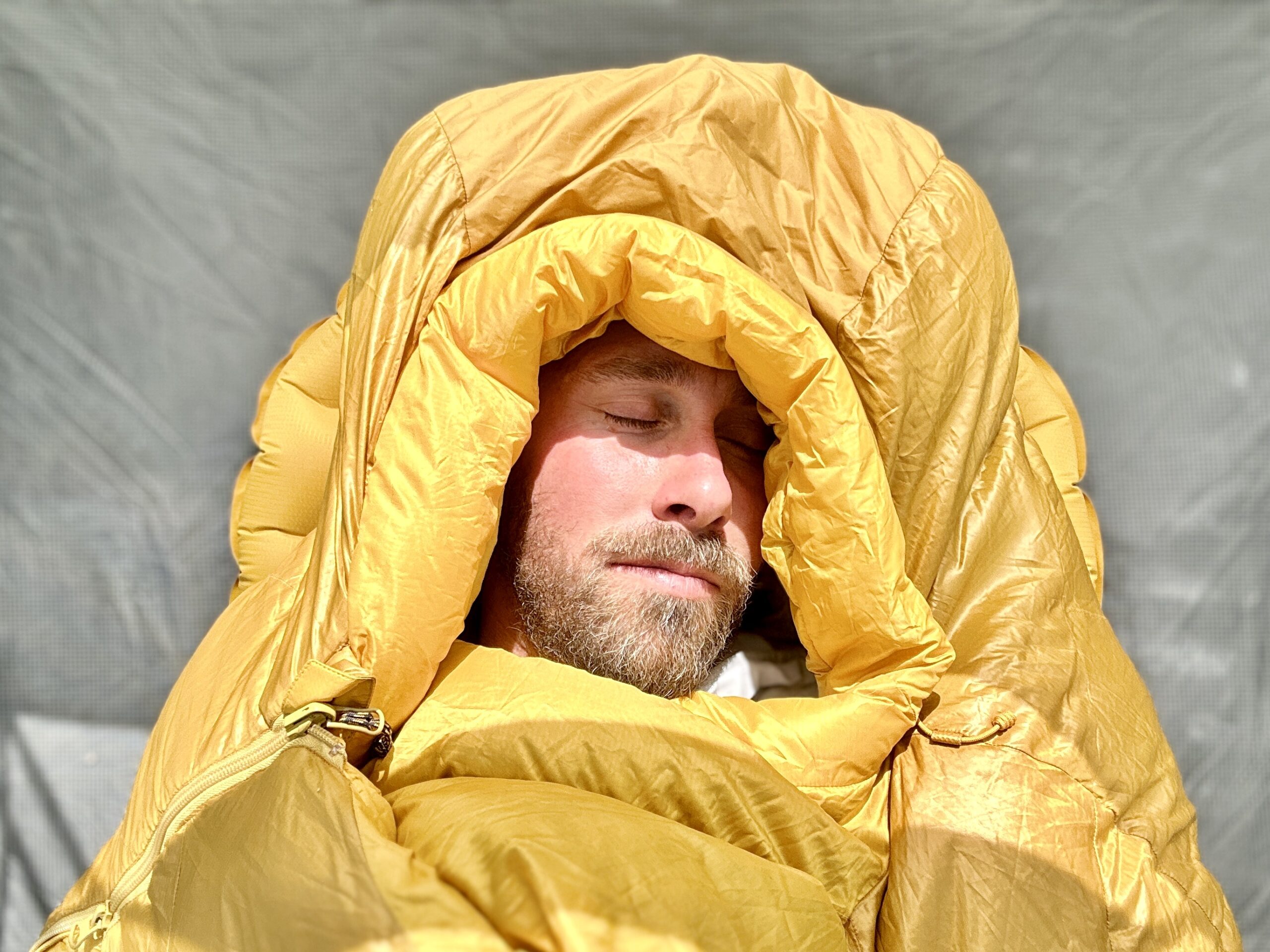 Aclose up image of a man sleeping in a sleeping bag, with morning sun on his face. The hood is cinched around his face and it looks very cushy and plush.