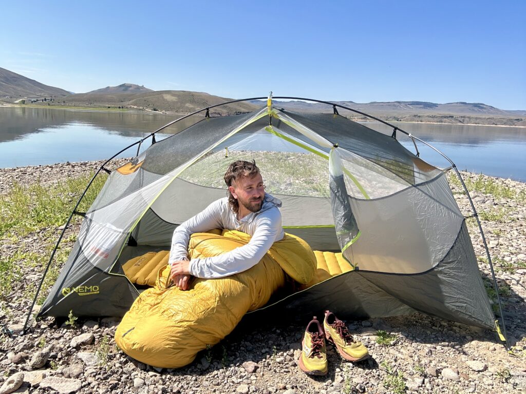 A man sits up in a sleeping bag with the footbox outside of his tent, looking off in the distance. His shoes are off to the side and there is a lake and low mountains behind him.