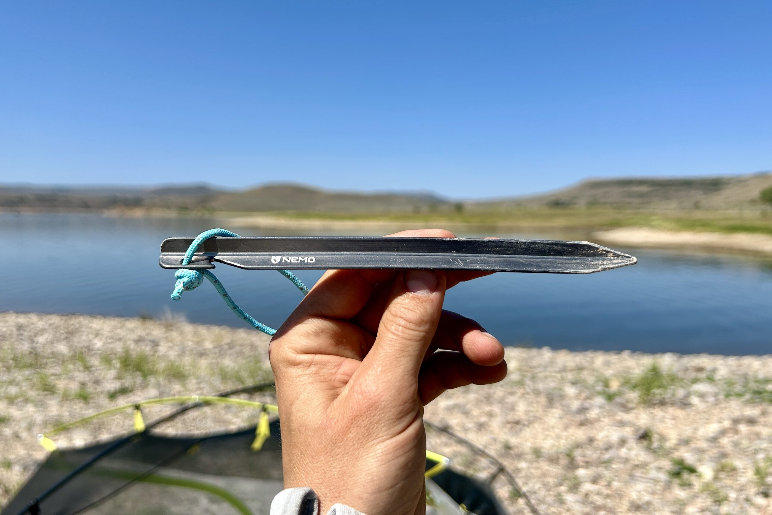 Close up of a hand holding a tent stake with water in background.