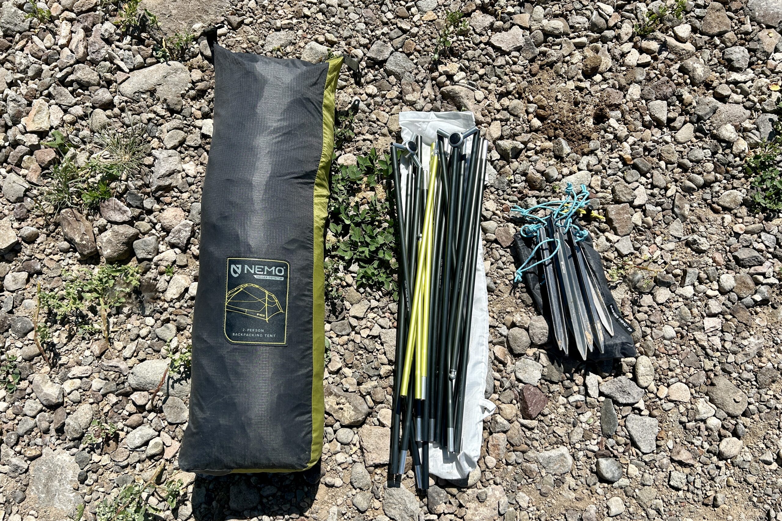 A tent stuff sack, poles, and stakes rest on the ground. next to each other.