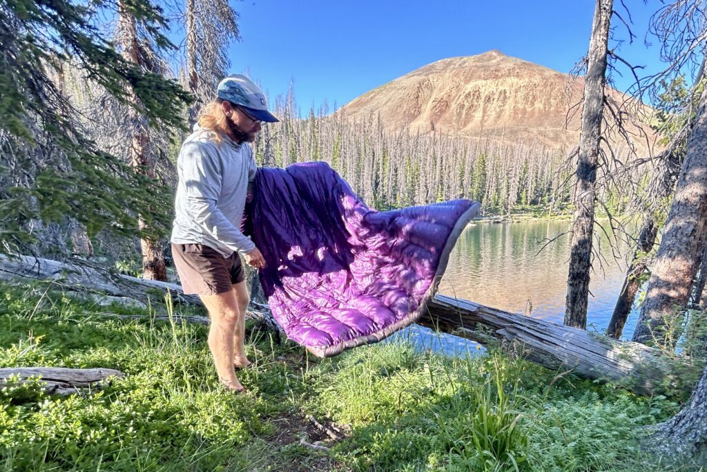 A man stands on the banks of a large alpine lake shaking out a purple quilt.