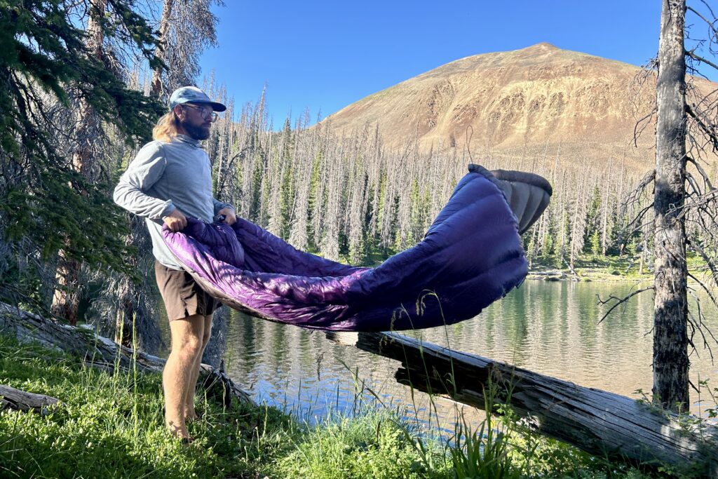 A man stands on the banks of a large alpine lake shaking out a purple quilt.