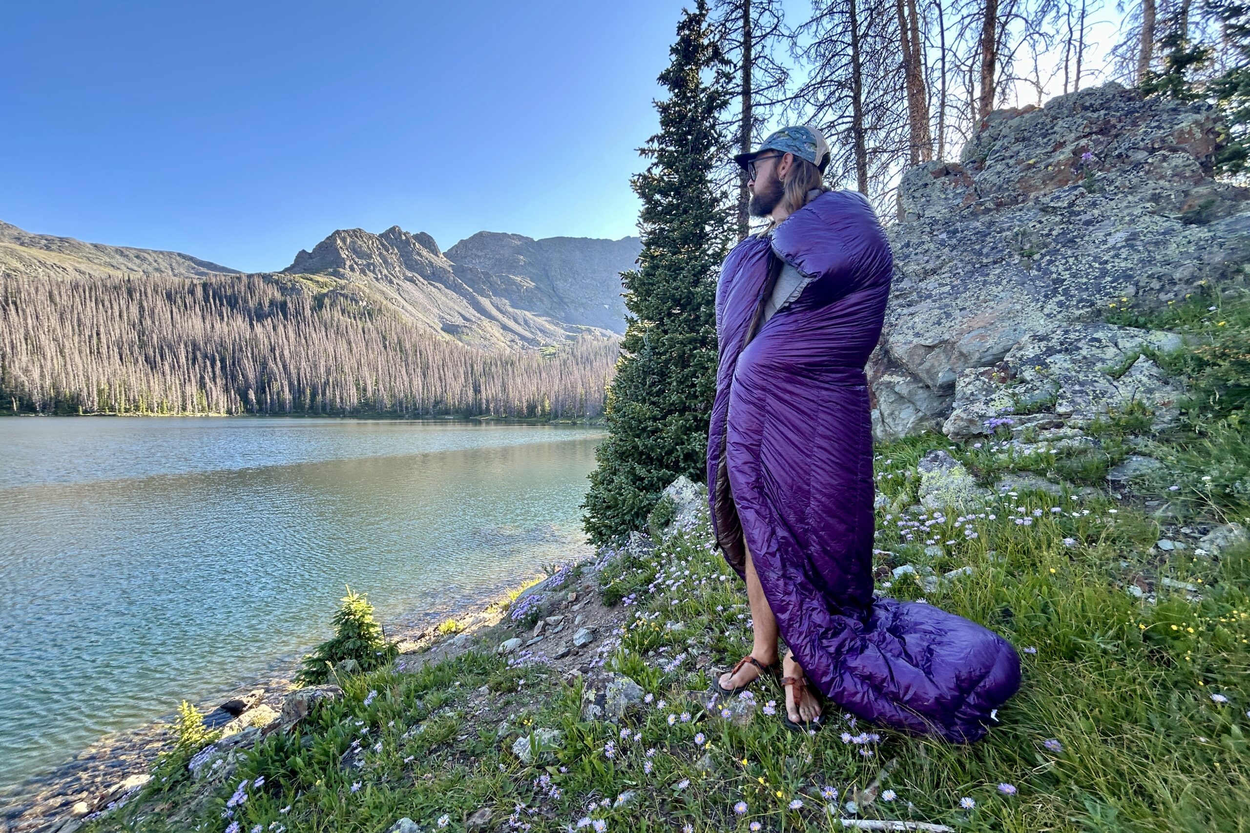 A man stands on the banks of a large alpine lake with a purple quilt wrapped around him. He is looking off in the distance towards mountains.