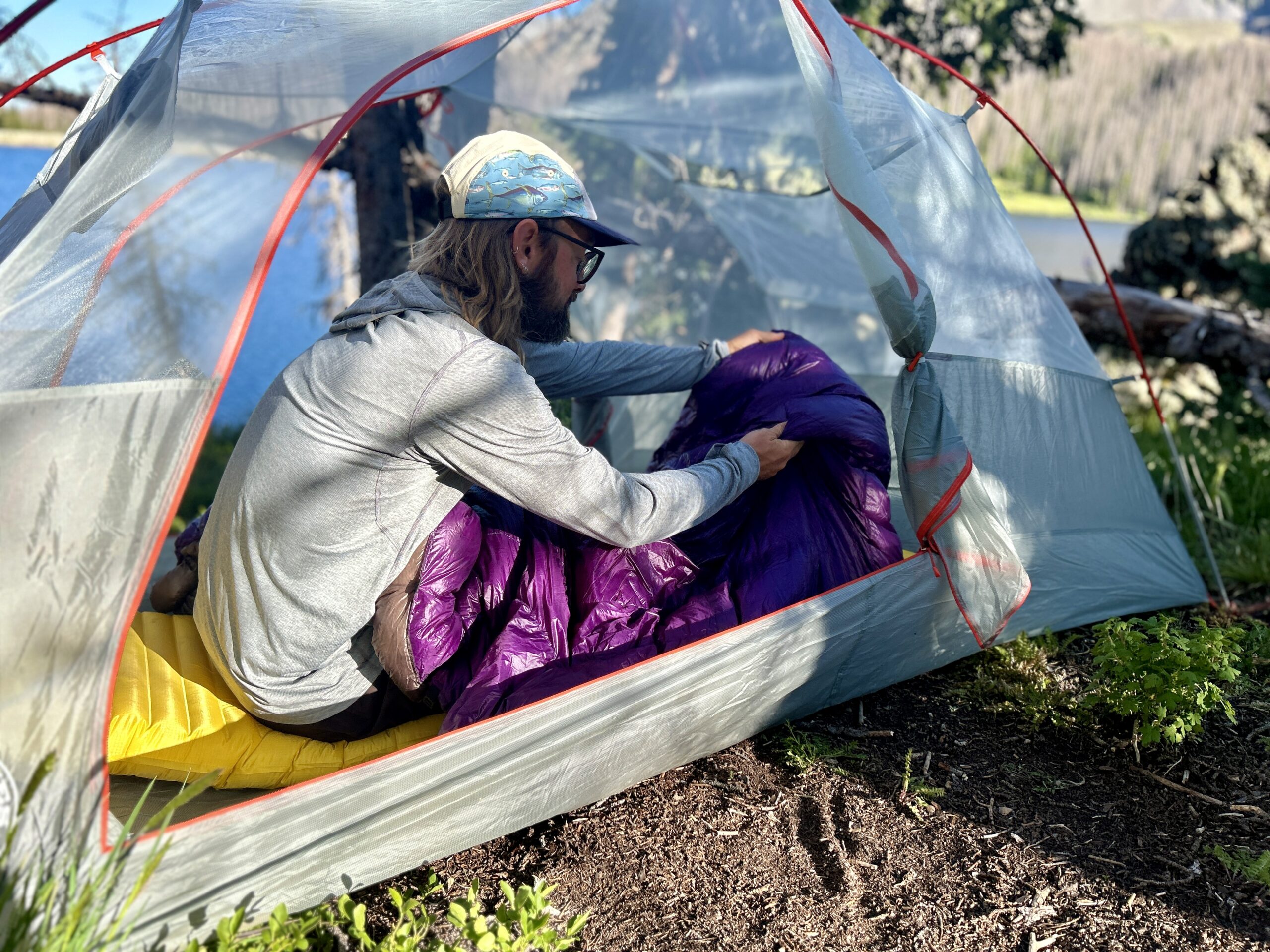 A man sitting in a tent with no rainfly adjusts his sleeping quilt while sitting up on a sleeping pad.