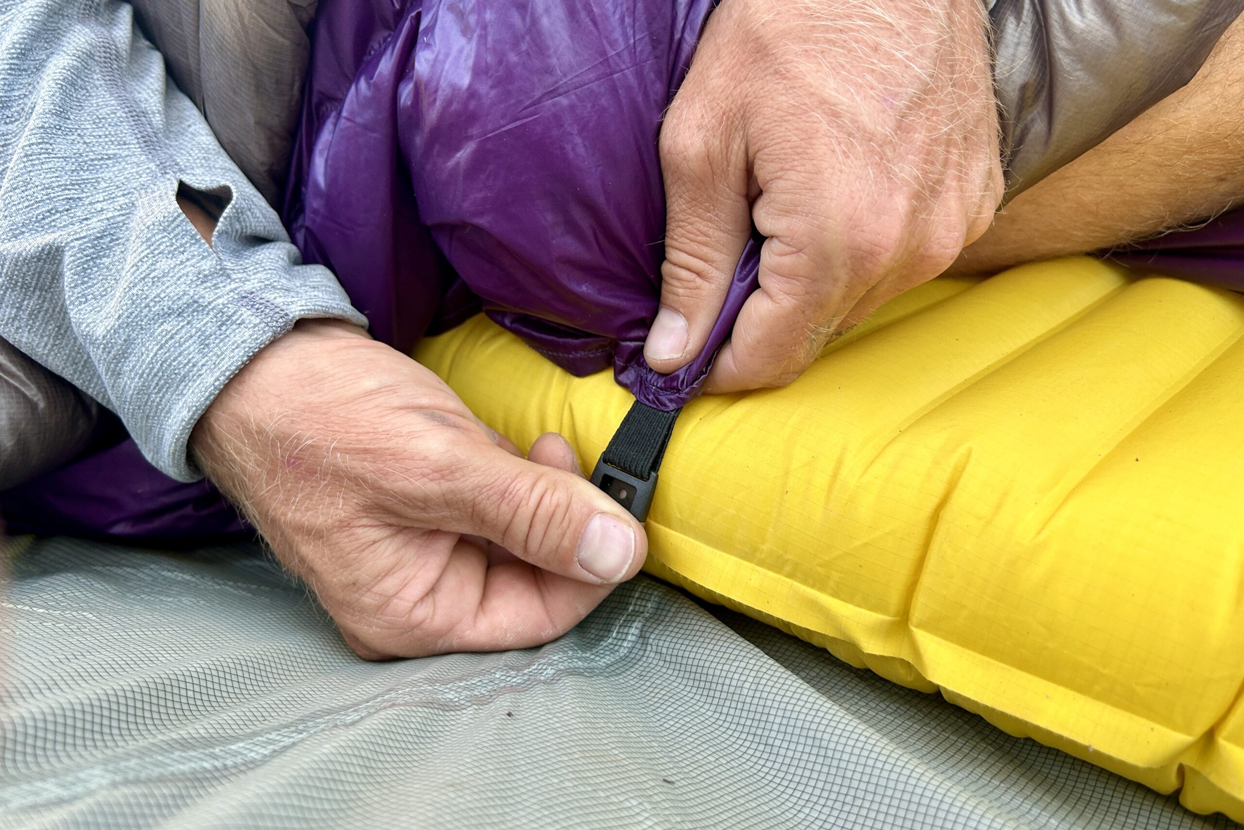 A person tightens the under-mattress attachment points of their quilt around their sleeping pad.