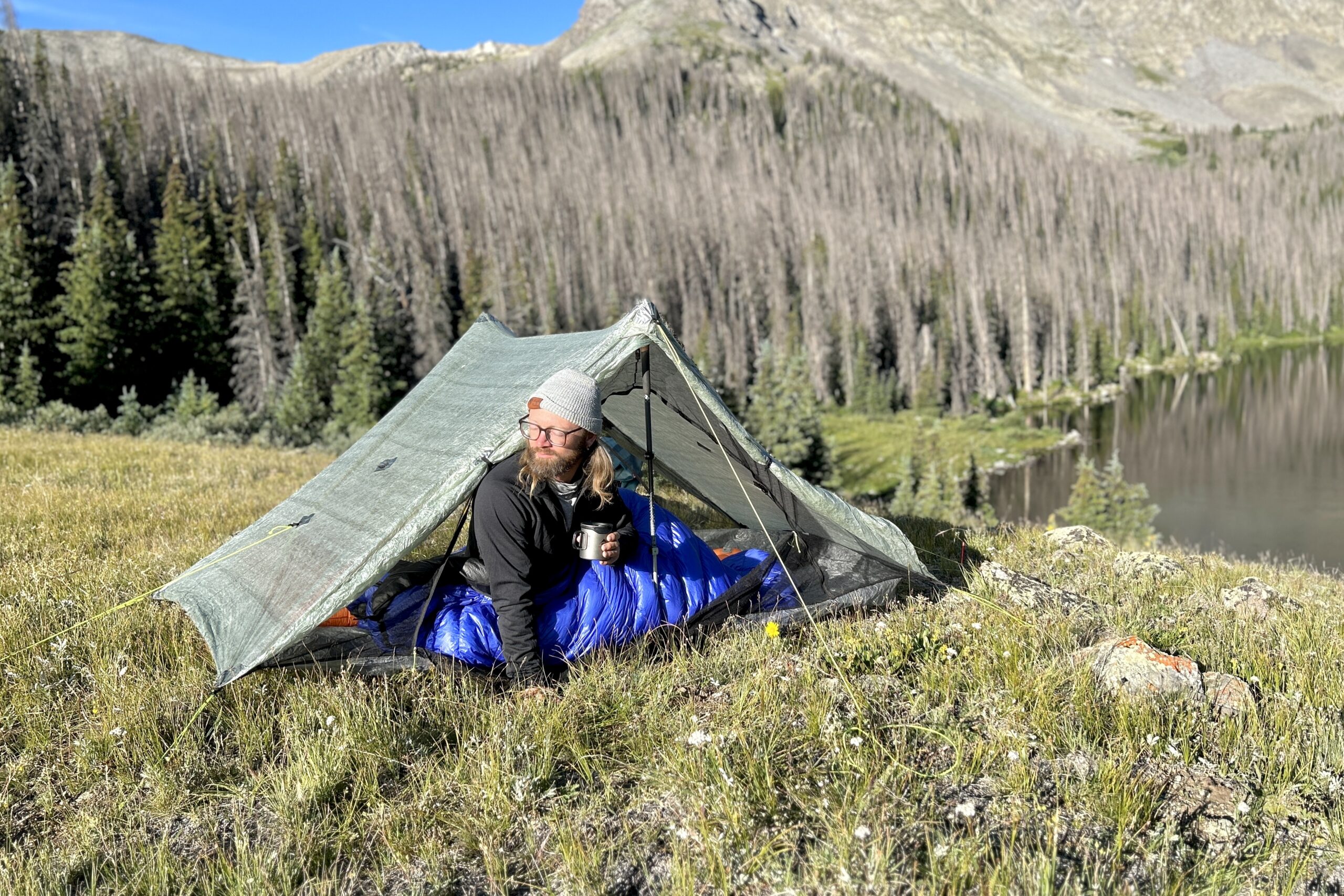 a backpacker holds their coffee or tea mug and sits up in their sleeping bag and sticks their head out of their zpacks duplex tent