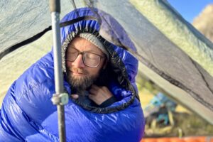 A man with glasses and a beanie on sits up in his tent. His eyes are closed.