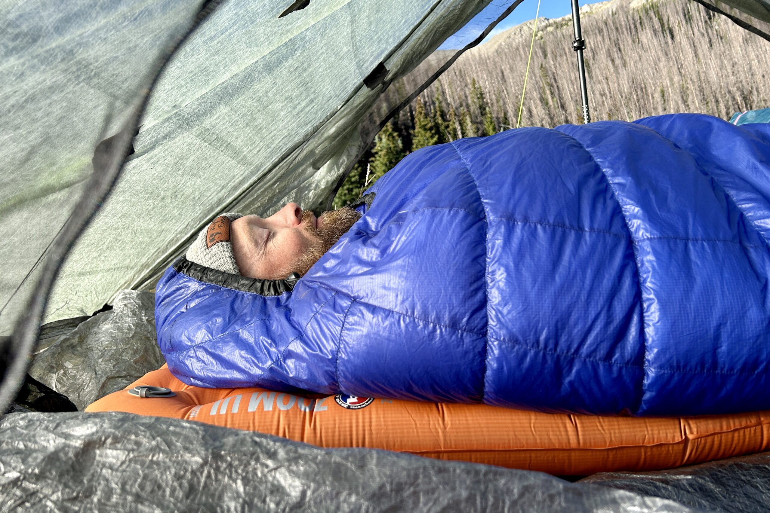 Visão de perto do perfil de um homem com um gorro dormindo em um saco de dormir todo fechado.