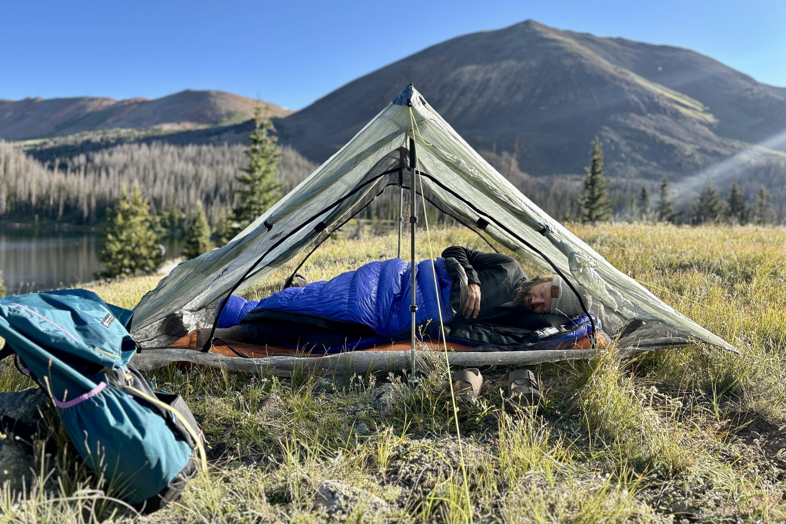 A man sleeping in a tent in a blue sleeping bag with his backpack on the ground next to his tent.