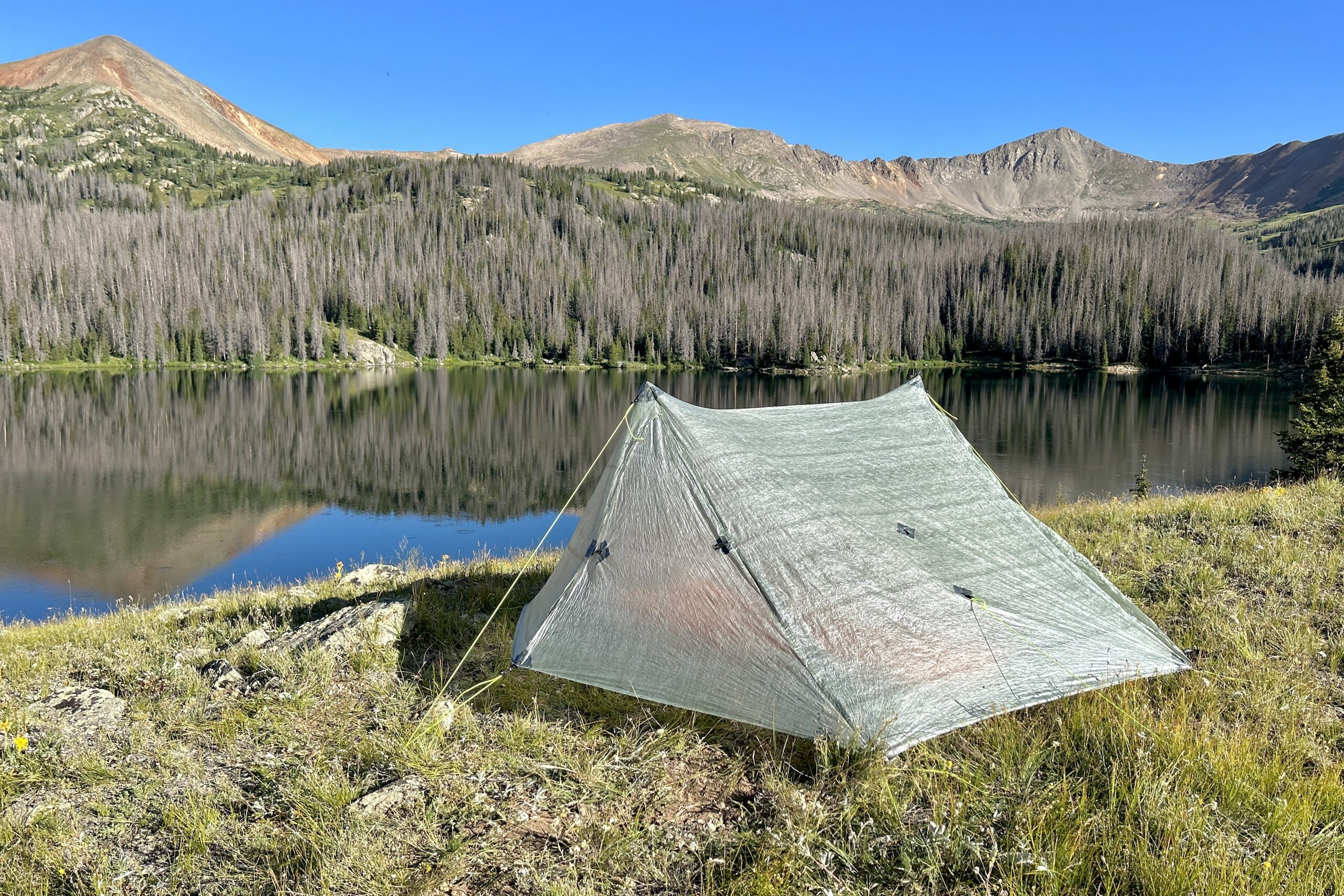 the zpacks duplex classic tent pitched near a glassy lake on a bright cloudless day. redish brown mountains enclose the lake