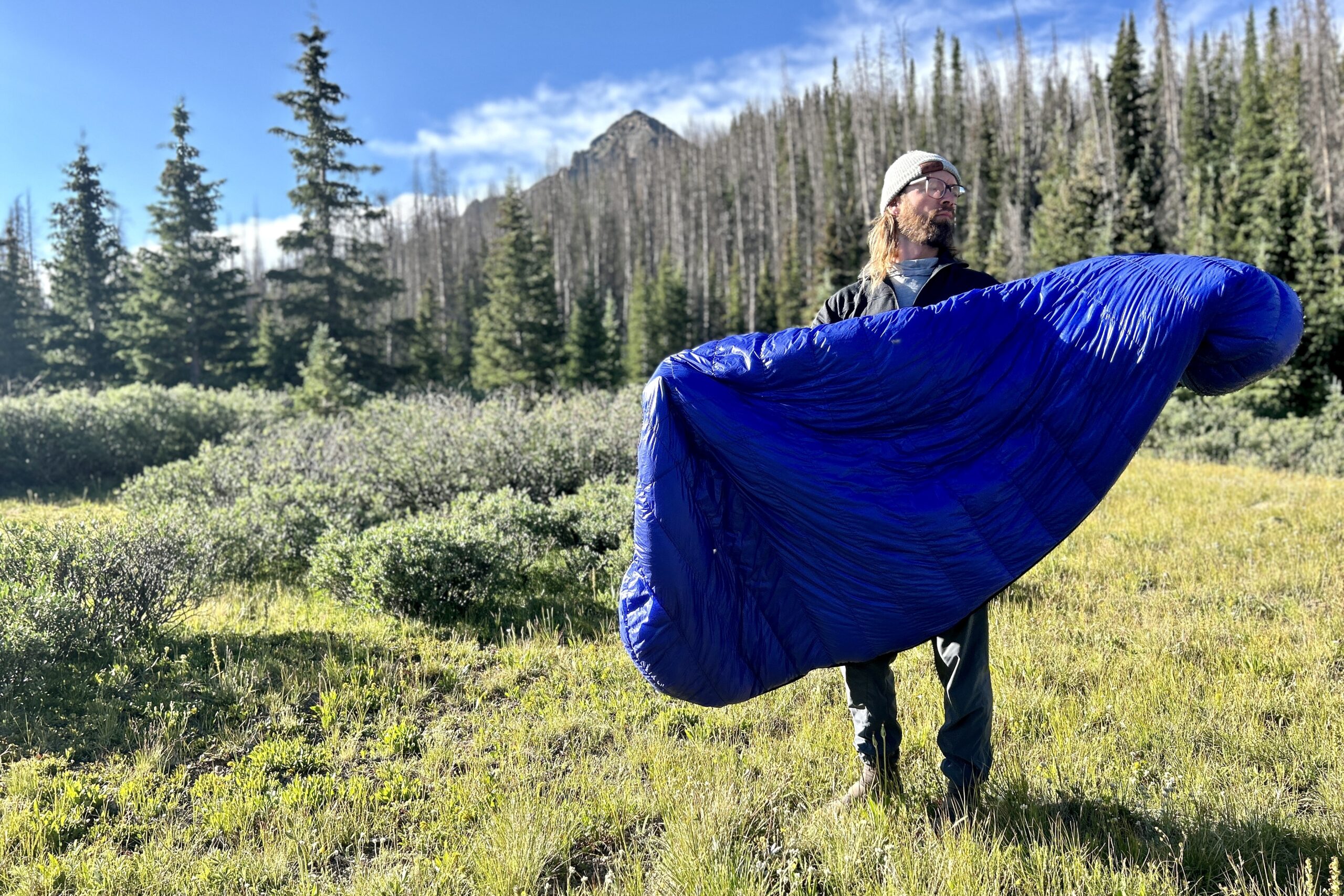 A man stands in field, folding a sleeping bag, next to tall grass and blue skies.