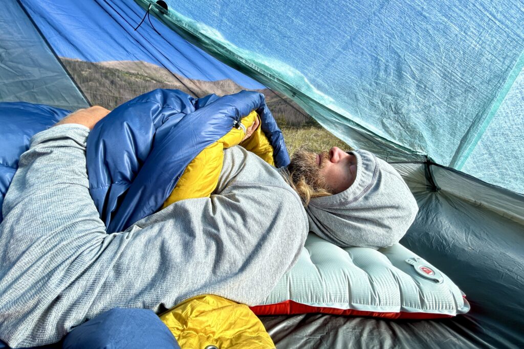 A profile image from the waist-up of a man with his hood up, eyes closed, and arms out and sleeping quilt pulled up underneath his arms.