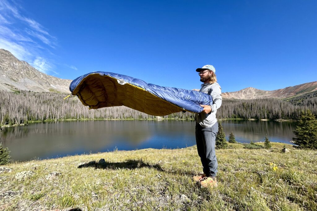 A man fluffs out a sleeping quilt in a field with a lake behind him.