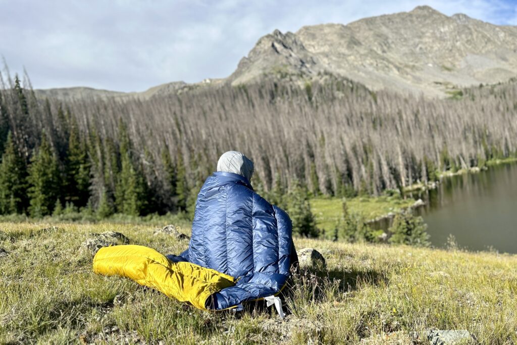 A man sits with a sleeping quilt wrapped around his back looking over an alpine lake with mountains behind it.