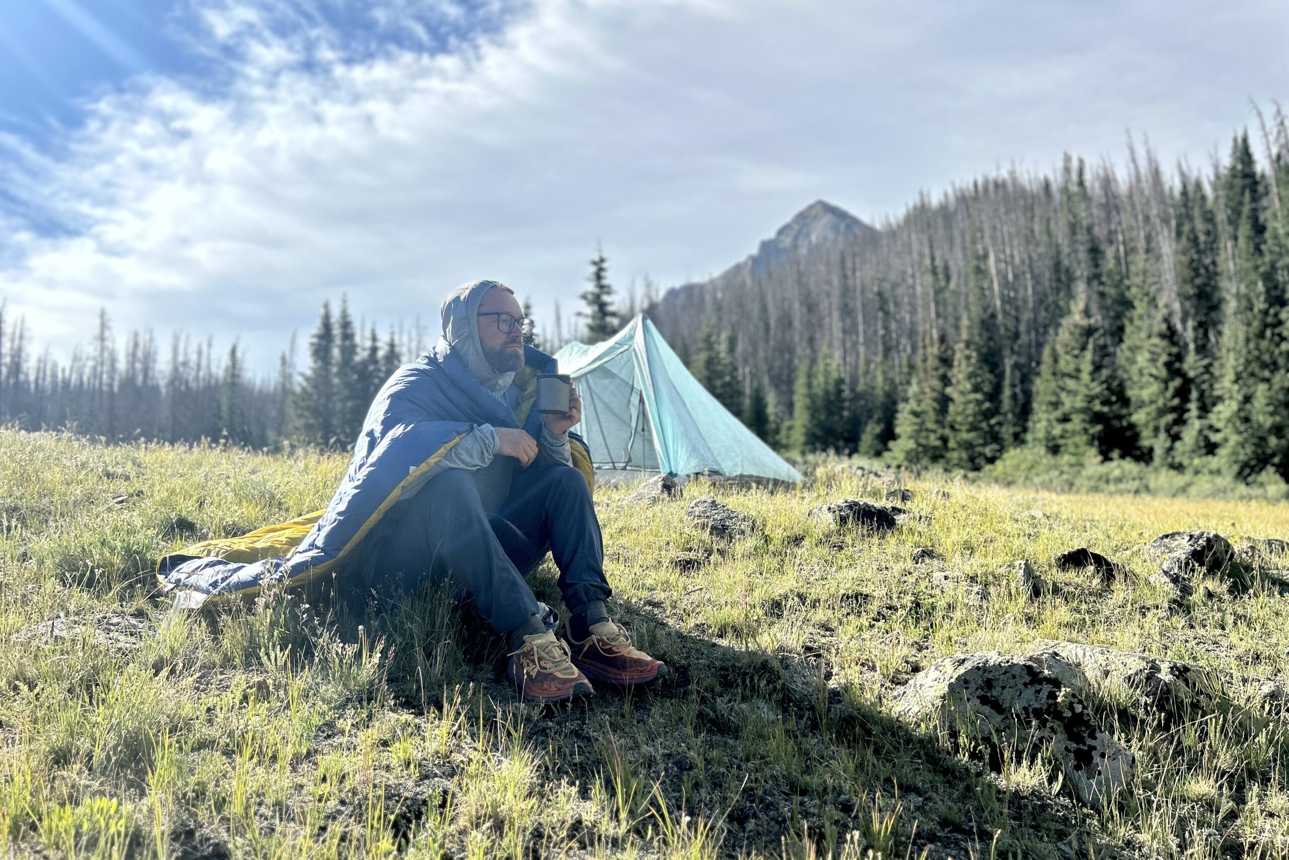 A man sits in a field of grass with his quilt wrapped around his back, sipping a coffee in front of his tent. It's a beautiful, sunny morning.