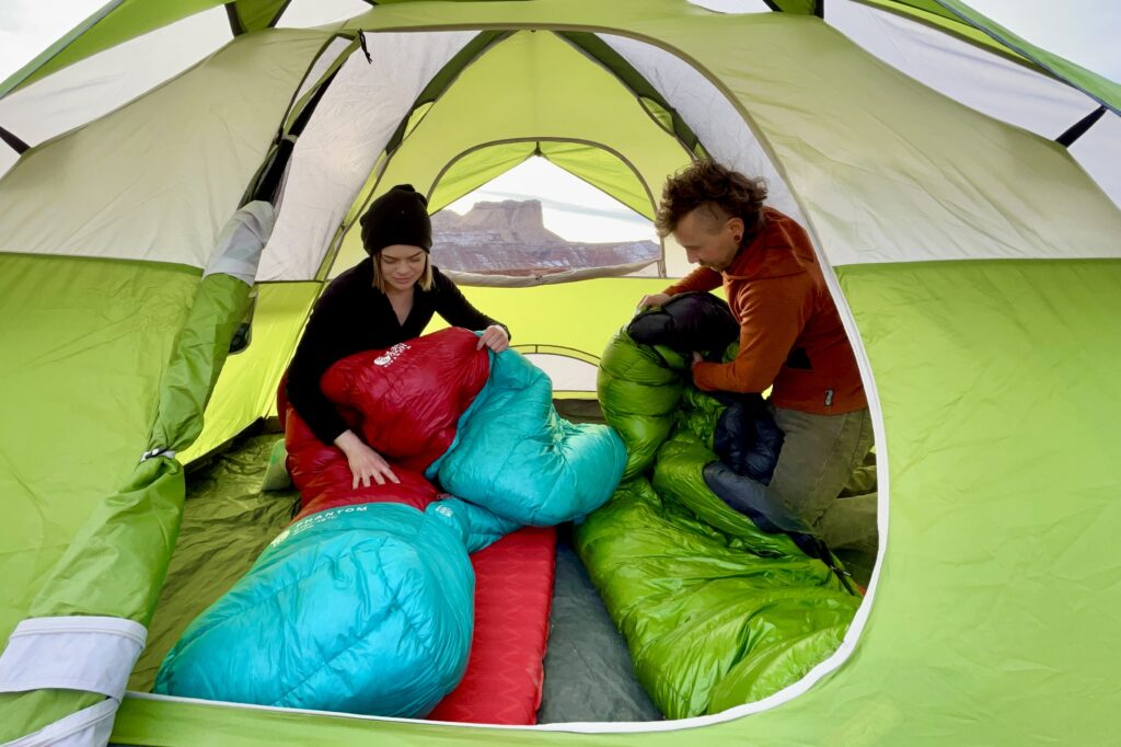 Two people pack up their sleeping bags in a green tent with a large desert butte visible in background.