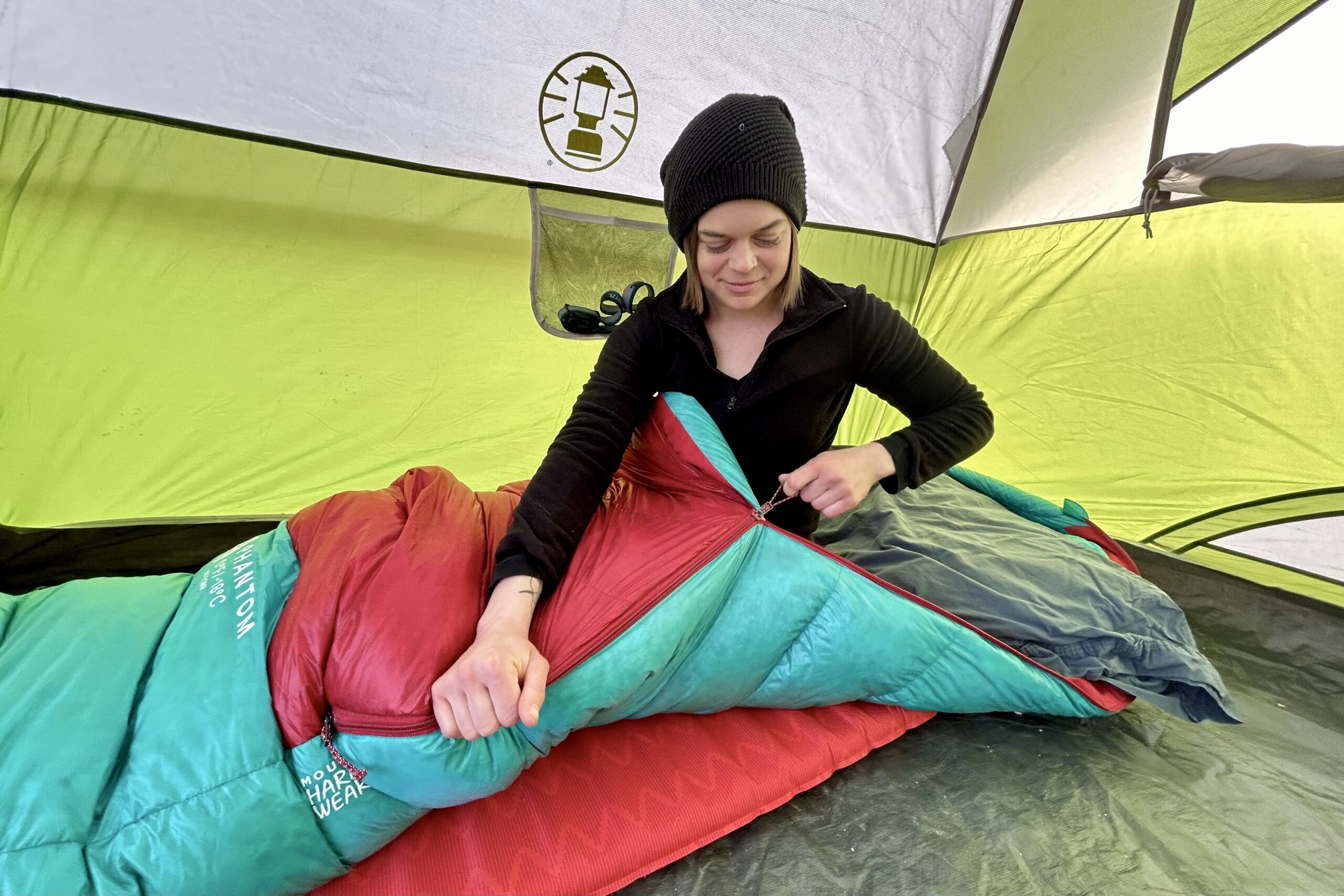 A woman wearing a beanie zips up a sleeping bag while sitting inside it.