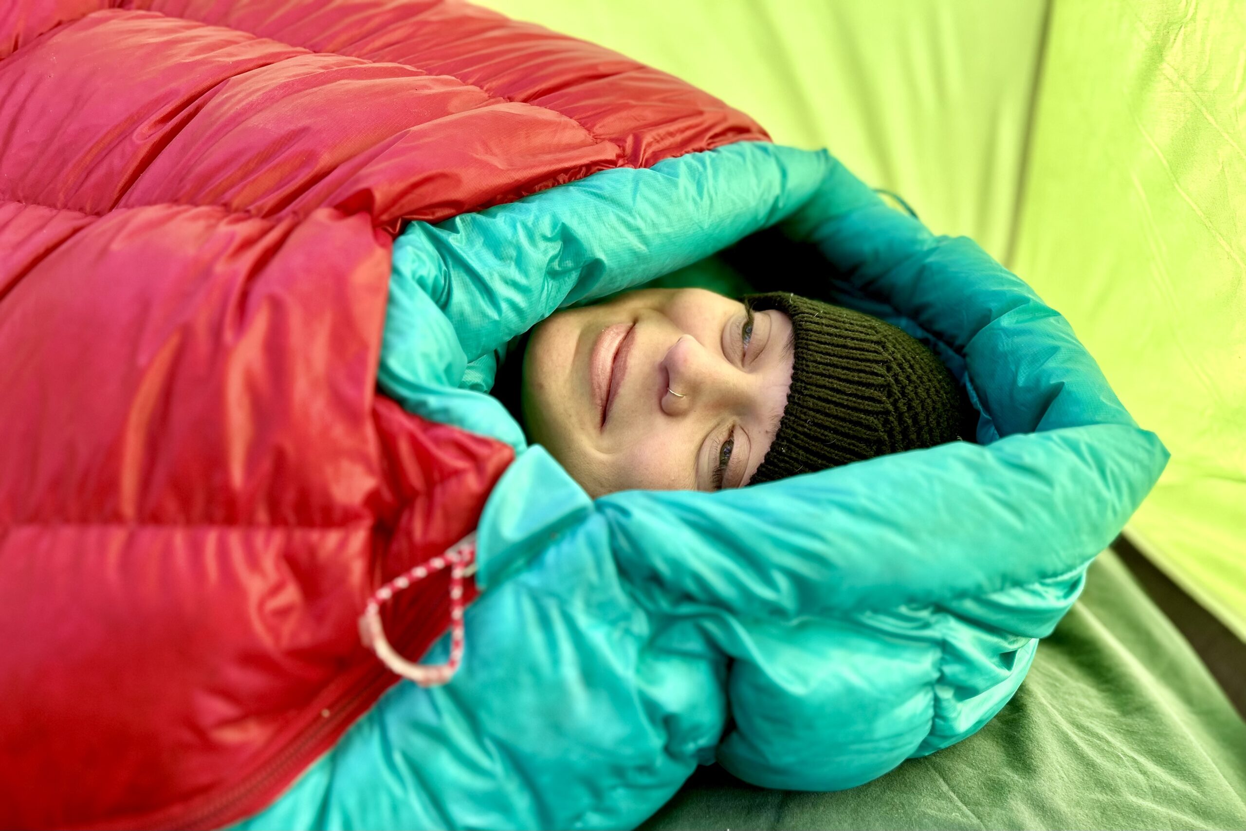 An up-close image of a woman smiling in a sleeping bag with the hood pulled up around her.