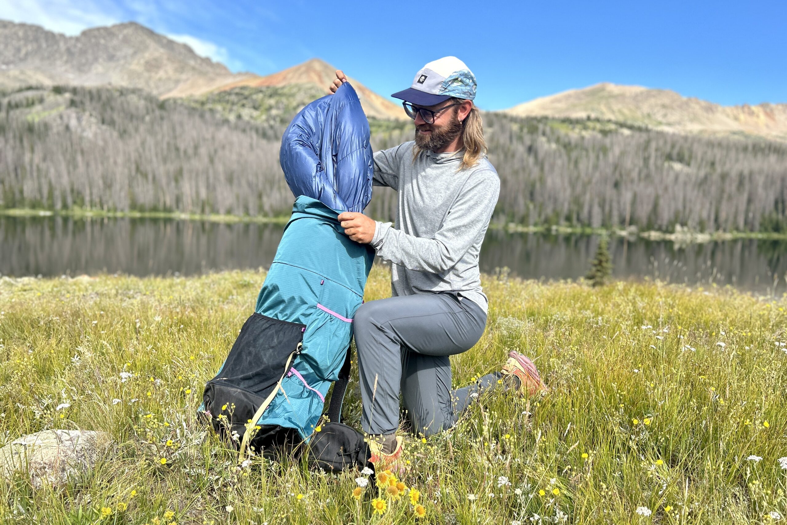 A man pulls a down quilt out of a backpack while kneeling in a field of flowers with a lake and mountains behind.