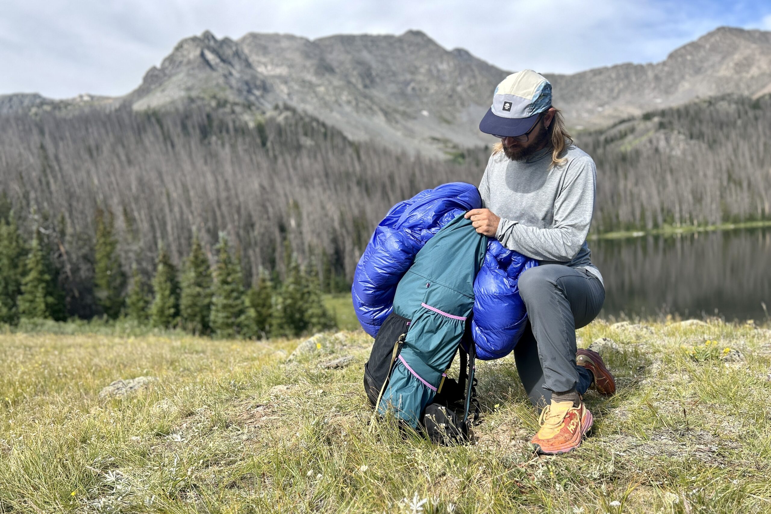 Um homem coloca um saco de dormir em uma mochila ao lado de um lago alpino com montanhas ao fundo.