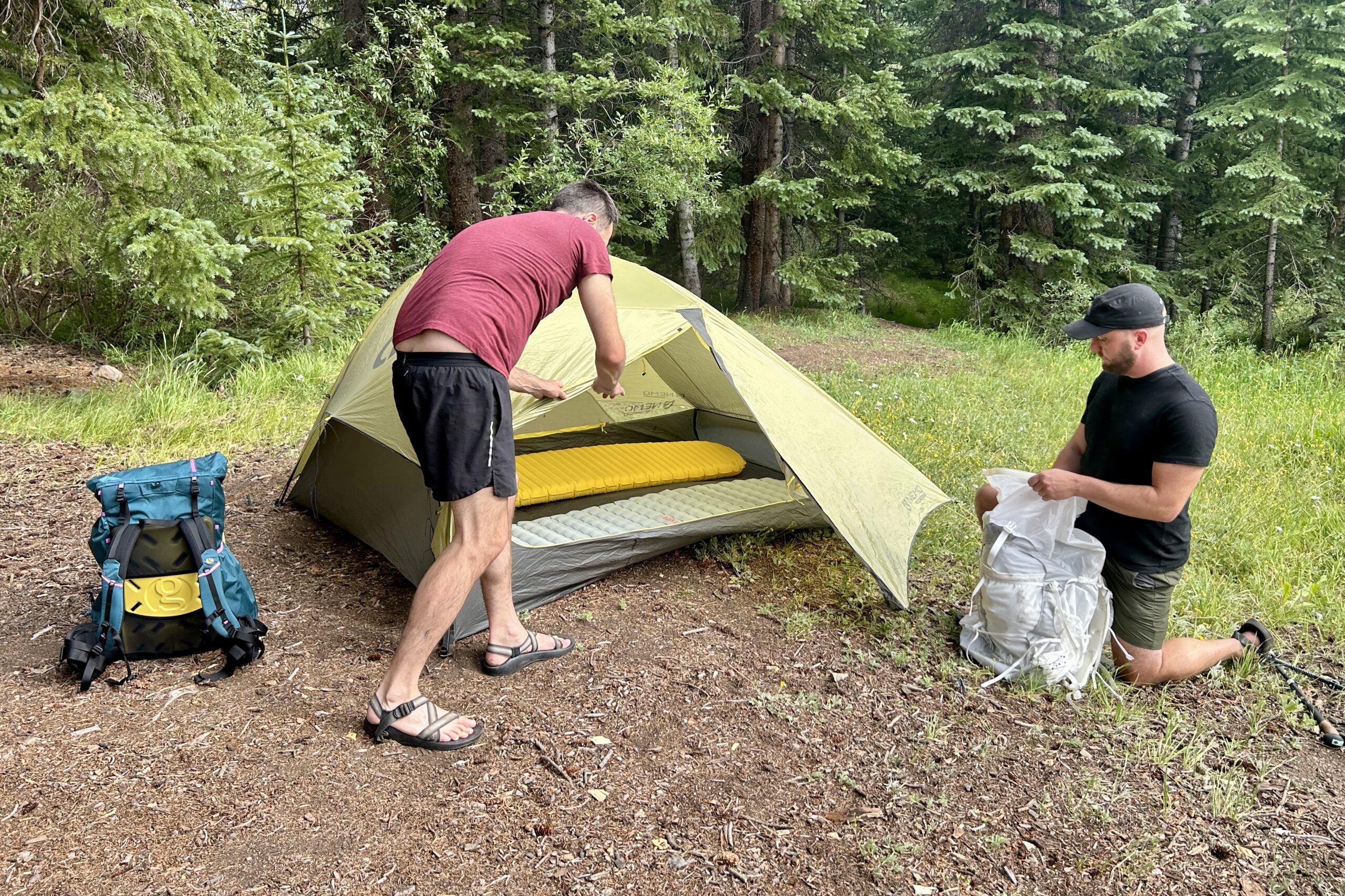 2 men in the woods unpack their backpacks and set up a tent with the rainfly.