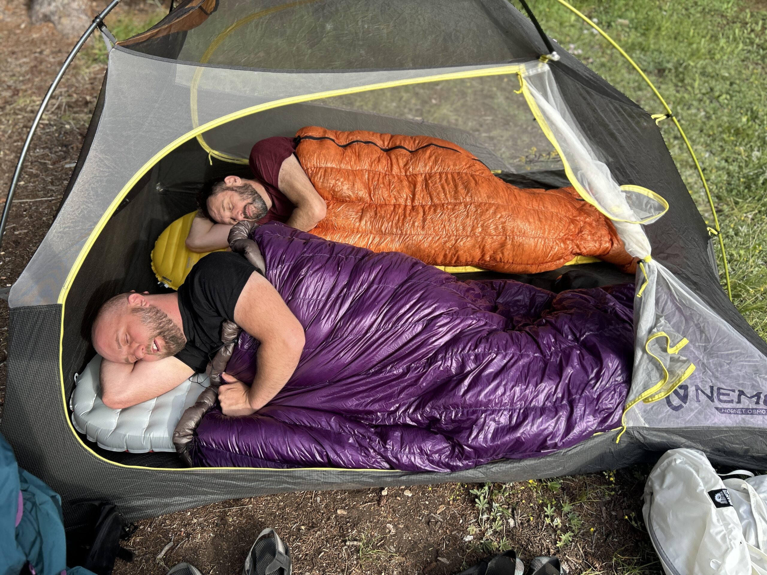 Two men sleep next two each other in an orange sleeping bag and a purple quilt in a 3-person tent. Both are facing towards the camera and using their arms as a pillow.