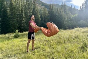 A man shakes out a sleeping bag in a grassy field on a hot summer day.