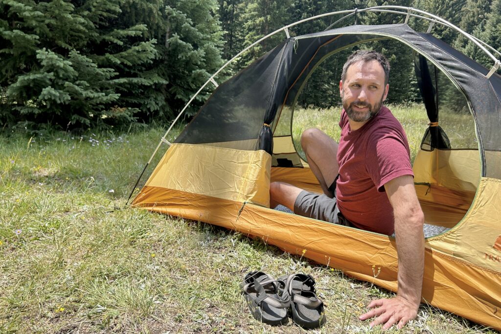 A man leans outside of a tent in the woods smiling.