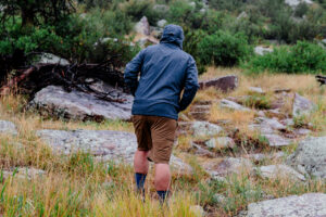 A man wearing a blue jacket and brown shorts hikes in the rain with shrubs and rocks in front of him.