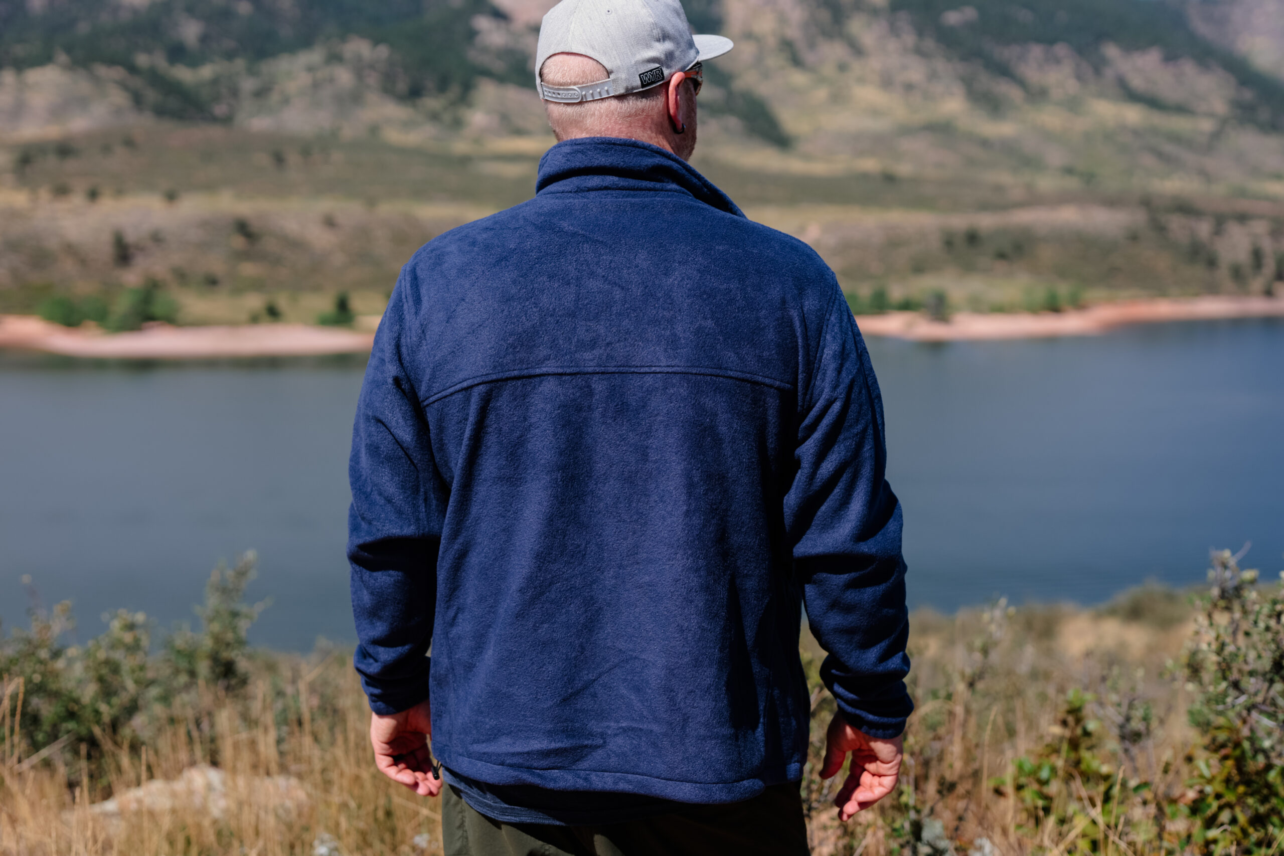 A man wearing a blue fleece's back as he faces a lake.