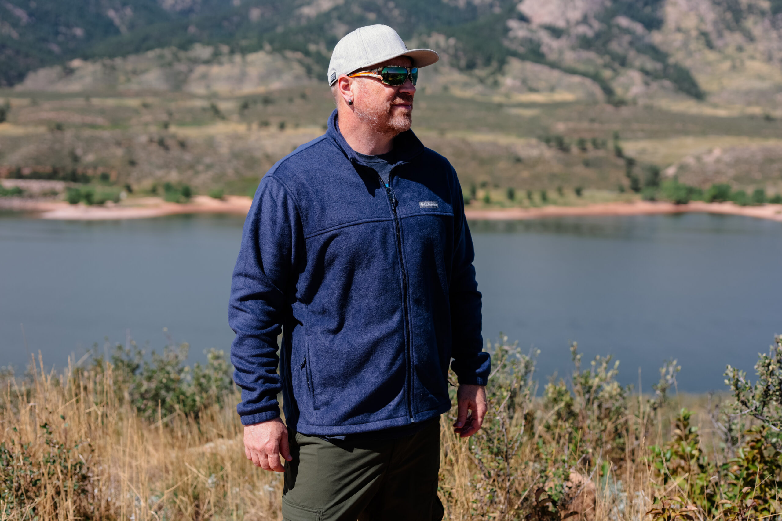 A man stands by a lake wearing a blue fleece.