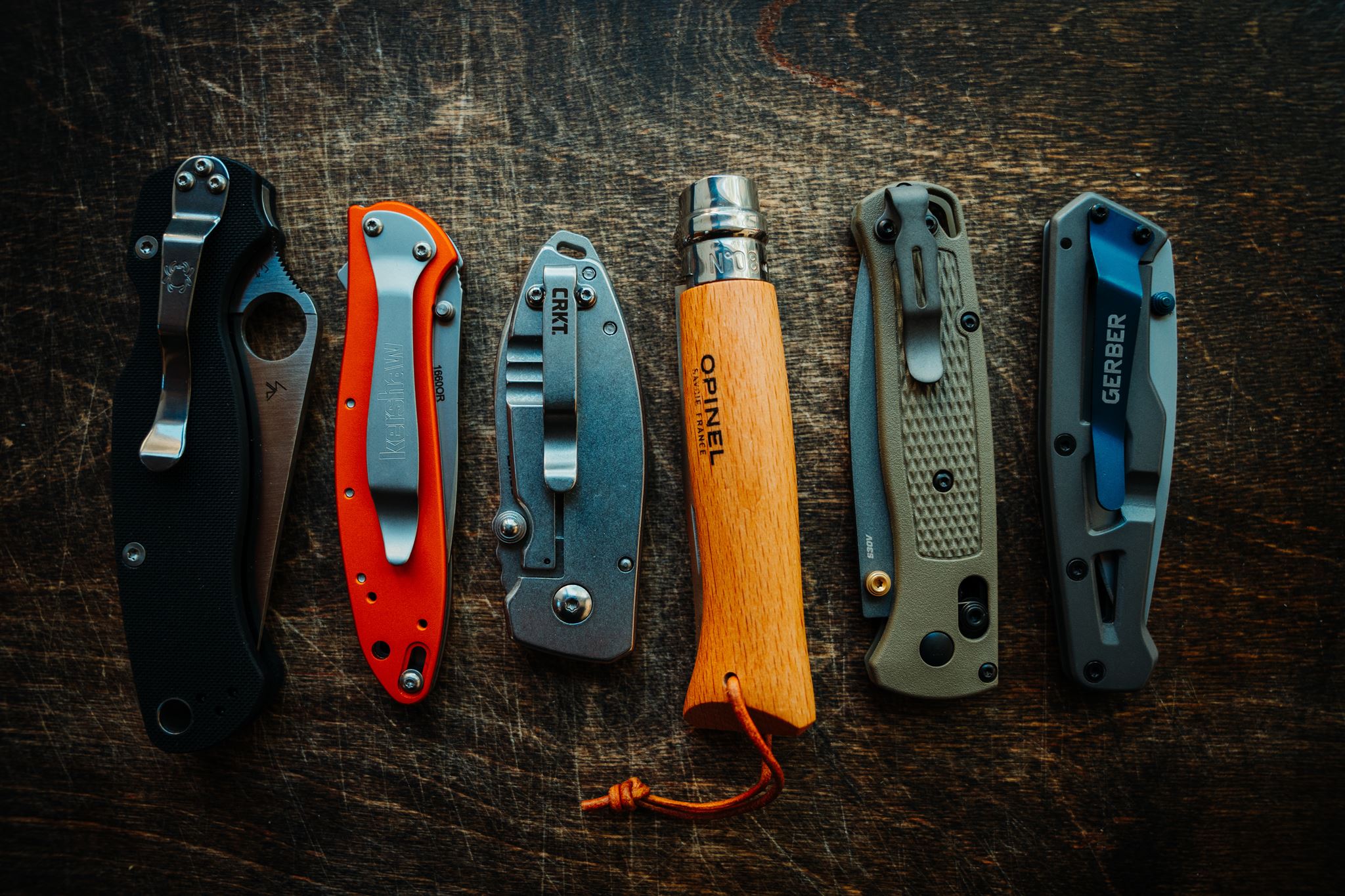 Six pocket knives are laid out side by side in the closed position to show size and build differences.