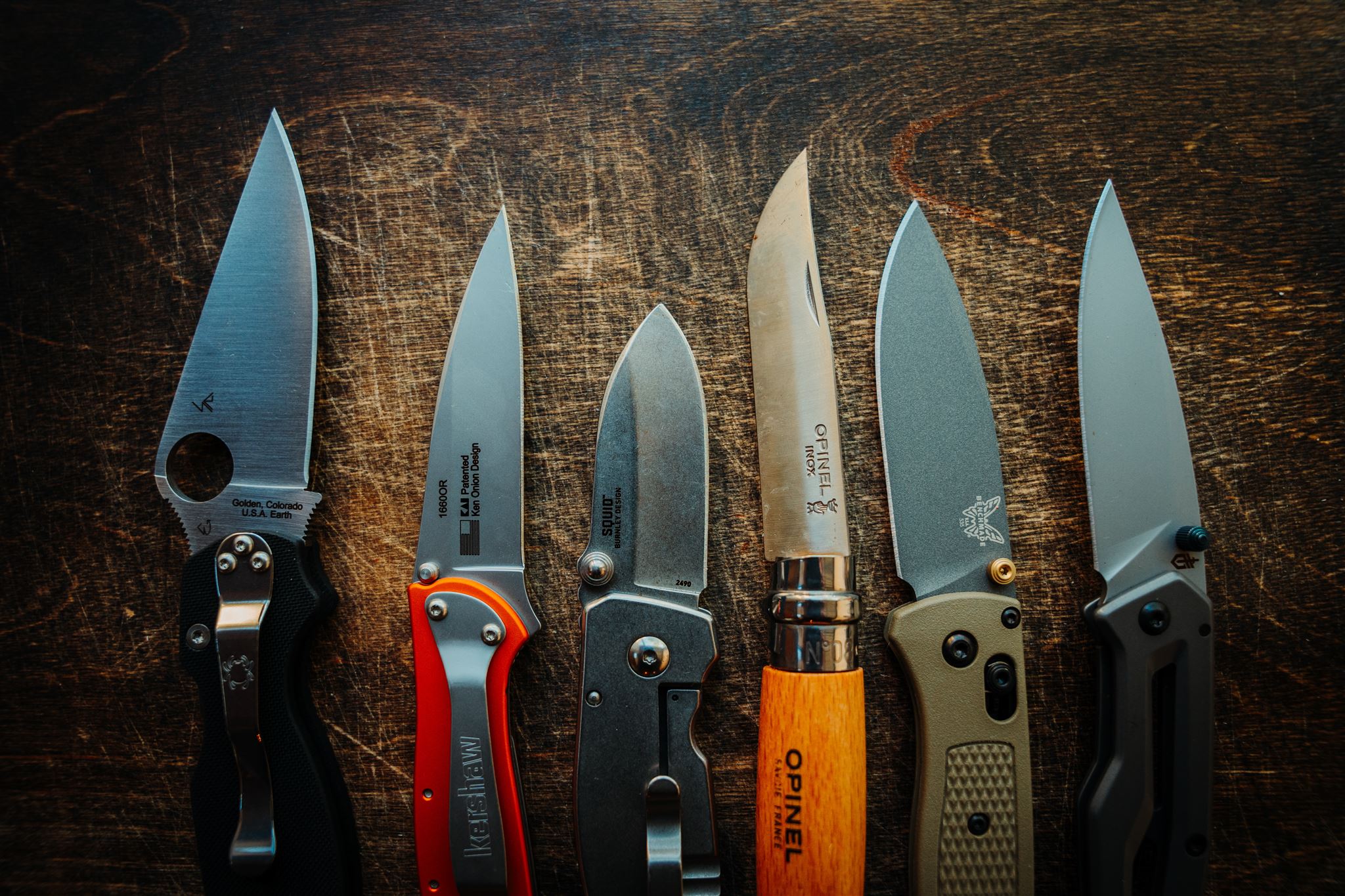 Six pocket knives are laid out on a wooden background with blades open to show blade length and shape.