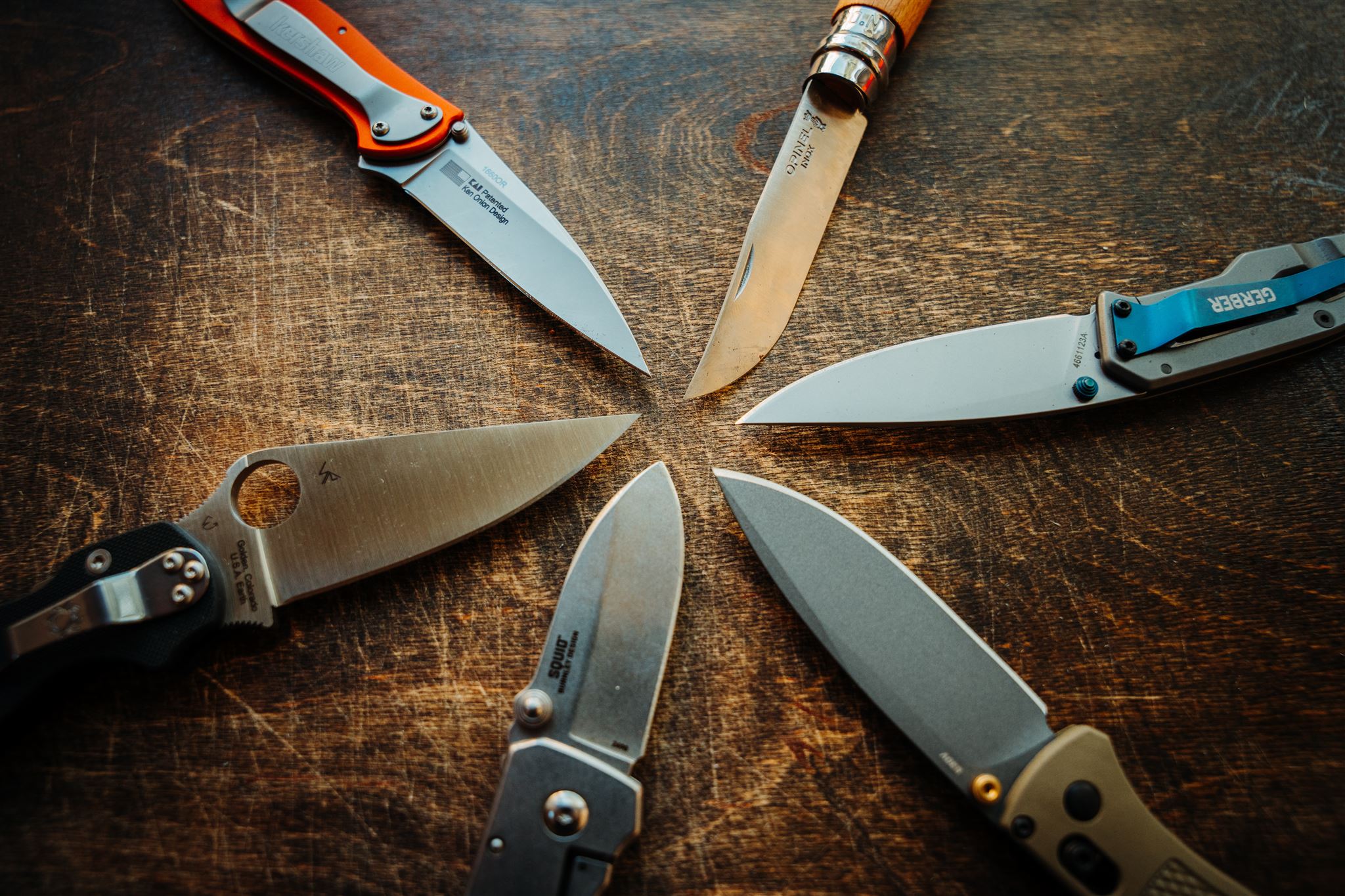 Six open pocket knives laid out in a circular manner to highlight the blade shape and points.