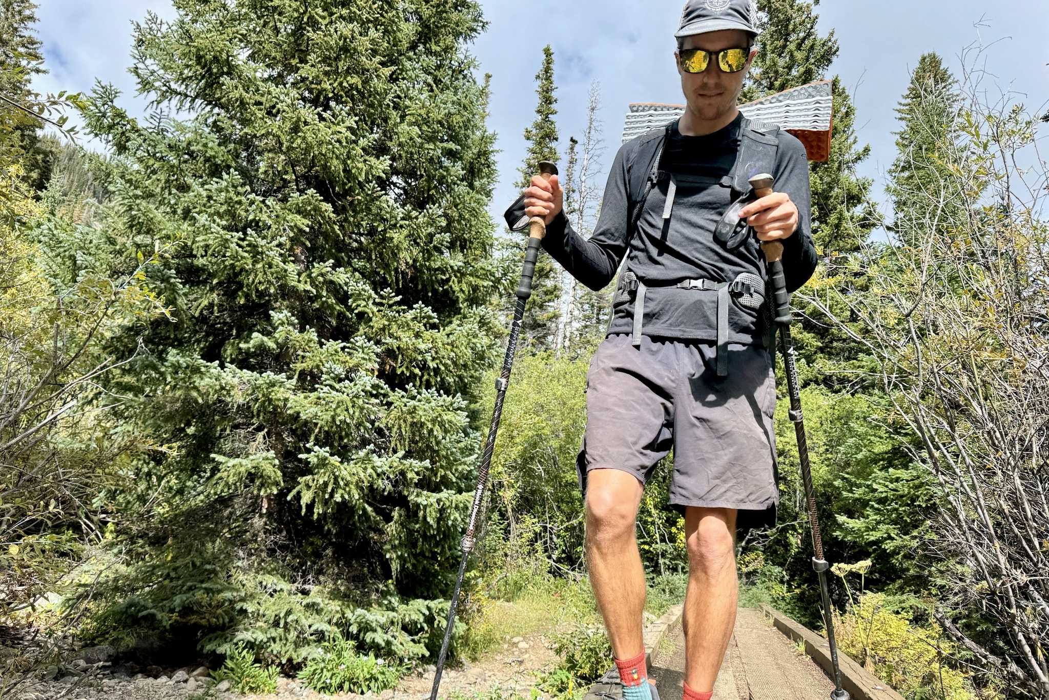 A hiker walks towards the camera with trekking poles, a pack, and wearing the Icebreaker Merino Oasis 200 base layer. Trees and a bridge are in the background.