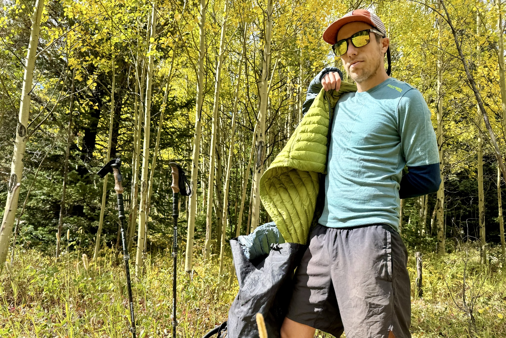 A hiker wearing the Ortovox Rock'n'Wool base layer while putting on a puffy jacket over it. Trekking poles and aspen trees are in the background.